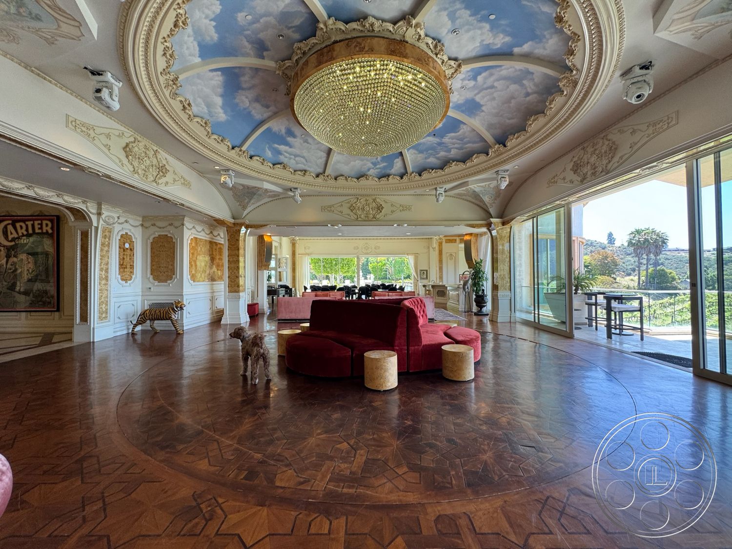 Beverly Compound - open floor plan, artwork on walls, glass sliding doors, dark hardwood flooring, decorative wall paneling, chandelier lighting fixture, intricate crown molding details, expansive windows with garden view, ornate ceiling with painted clouds, round parquet floor design, red velvet circular sofa, luxurious living area, stylized furniture decor