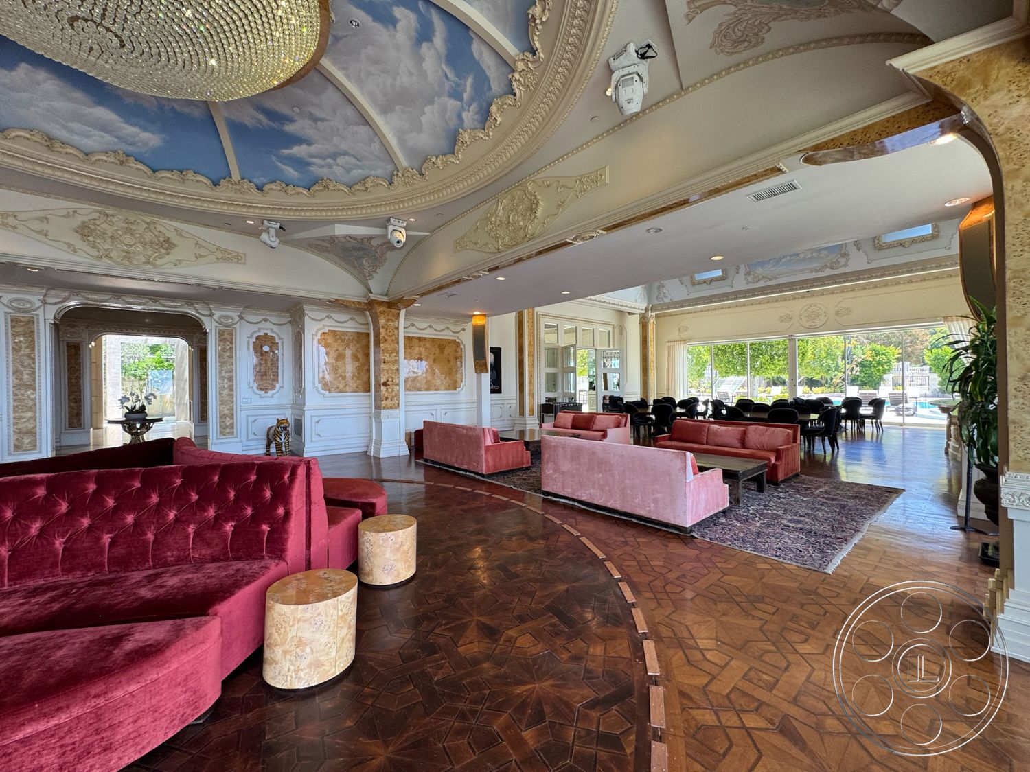 Beverly Compound - chandelier lighting, textured wallpaper, wood parquet flooring, plush velvet seating, open floor layout, large windows with outdoor view, rounded sectional sofa, coffered ceiling with decorative molding, gold-accented wall panels, circular accent tables, dining area with black upholstered chairs, elegant decorative elements, opulent design style