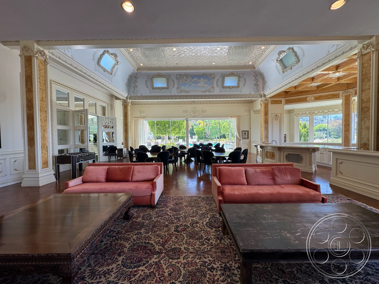 Beverly Compound - hardwood flooring, exposed wooden ceiling, elegant light fixtures, dark wood coffee table, marble bar countertop, intricate ceiling moldings, large windows with drapery, pink velvet sofa, ornate wall trims, dining area with circular tables, decorative ceiling medallions, classic area rug