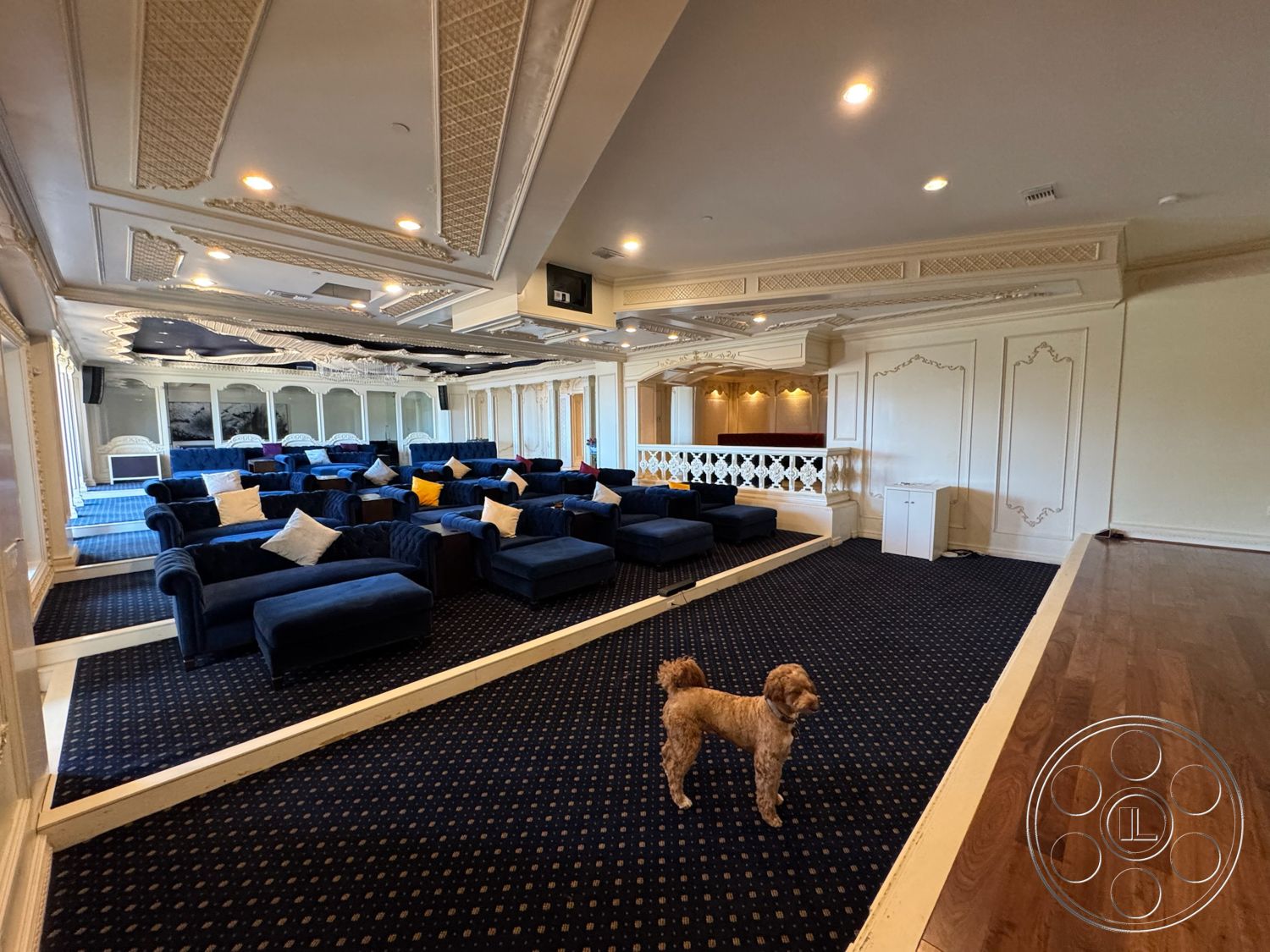 Beverly Compound - dark hardwood flooring, recessed lighting fixtures, textured wallpaper accent wall, wrought iron balcony railing, built-in sofa seating, plush blue velvet sofas, white decorative ceiling moldings, navy patterned carpet flooring, multi-tiered ceiling design, art deco style decor