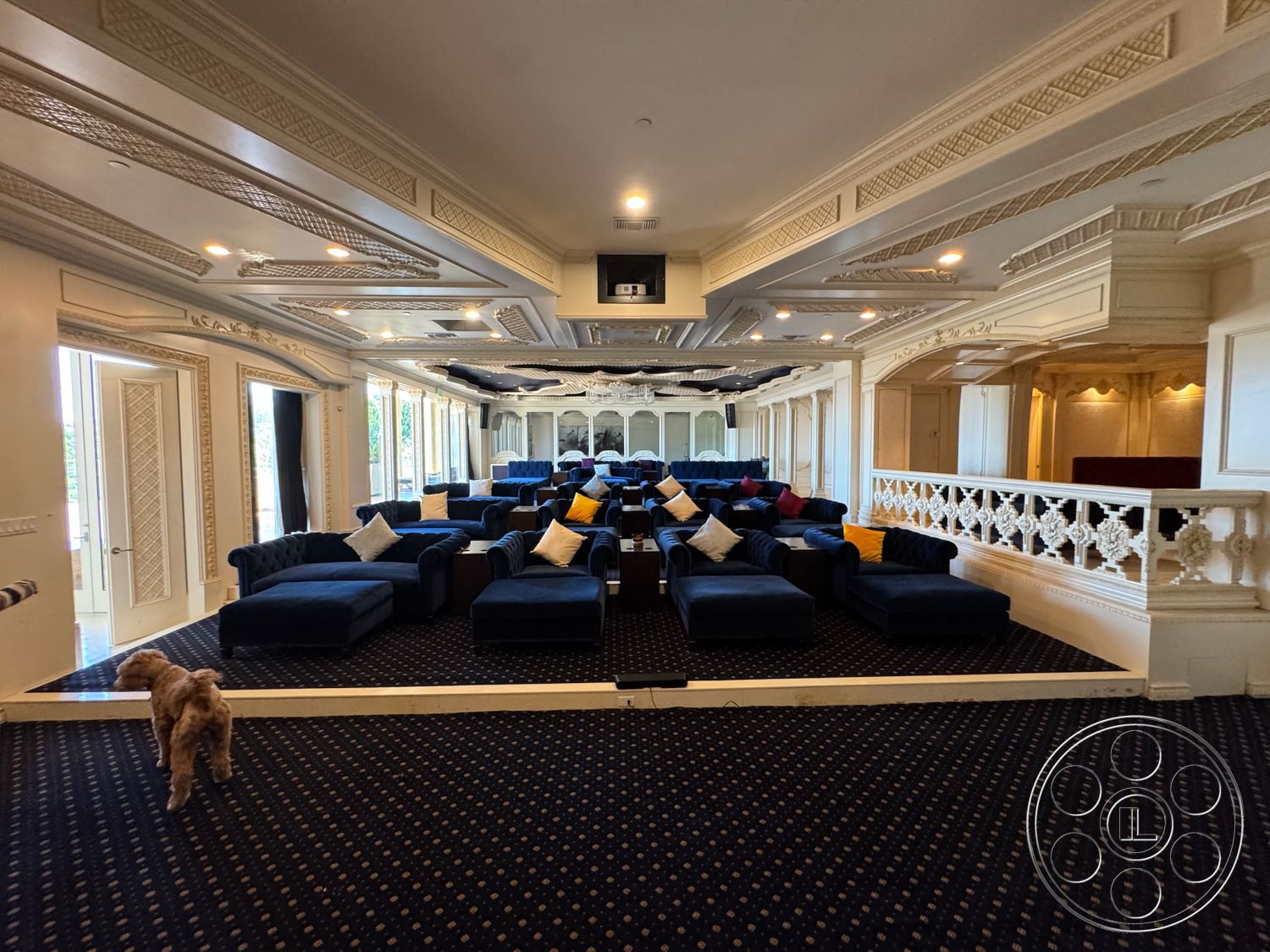 Beverly Compound - dimmed recessed lighting, double door entrance, intricate coffered ceiling, luxurious home theater room, plush blue velvet seating, gold leaf decorative moldings, detailed wall paneling, dark patterned carpet flooring, balustrade with ornate detailing