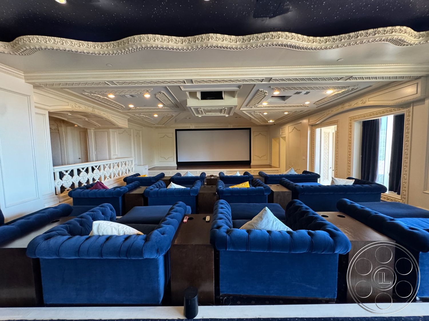 Beverly Compound - carpet flooring, decorative crown molding, projector screen wall, white paneled walls, home theater room, coffered ceiling with ornate details, starry sky ceiling, navy blue tufted velvet sofas, dark wood coffee tables, floor-to-ceiling windows with dark drapes