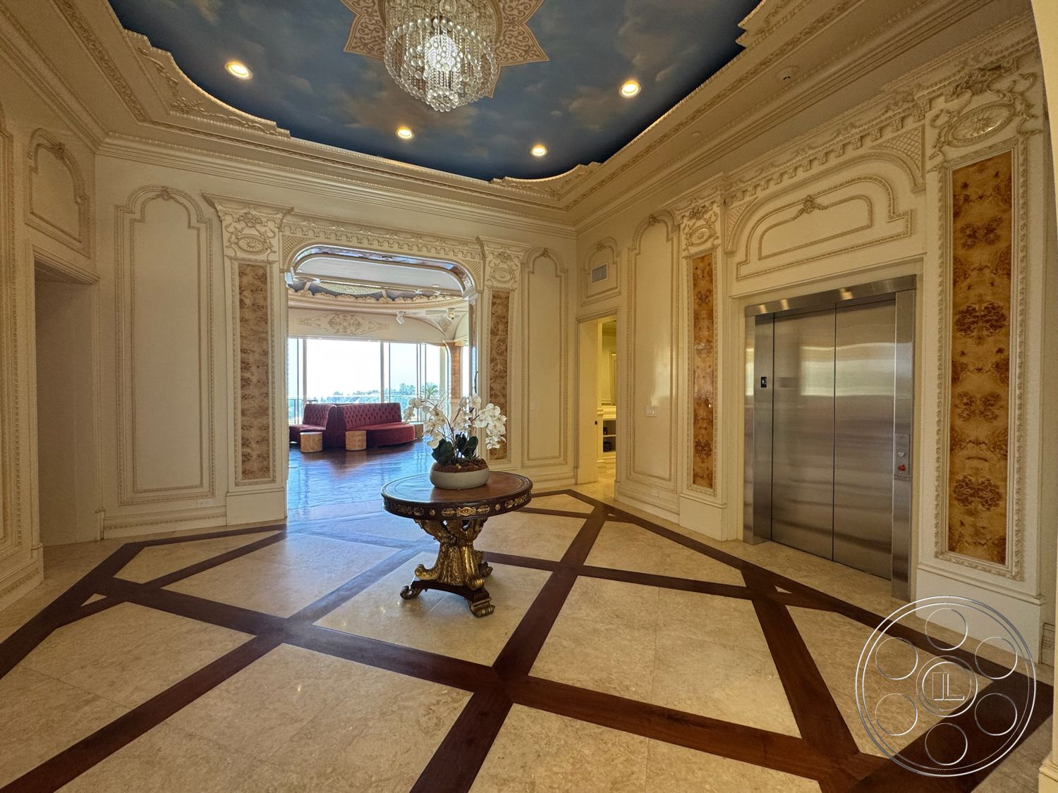 Beverly Compound - arched doorways, textured plaster walls, elevator access, contemporary seating area, decorative columns, chandelier light fixture, golden accents, large windows with city view, coffered ceiling with sky mural, marble flooring with wood inlay pattern, ornamental wall moldings, exposed hardwood trim
