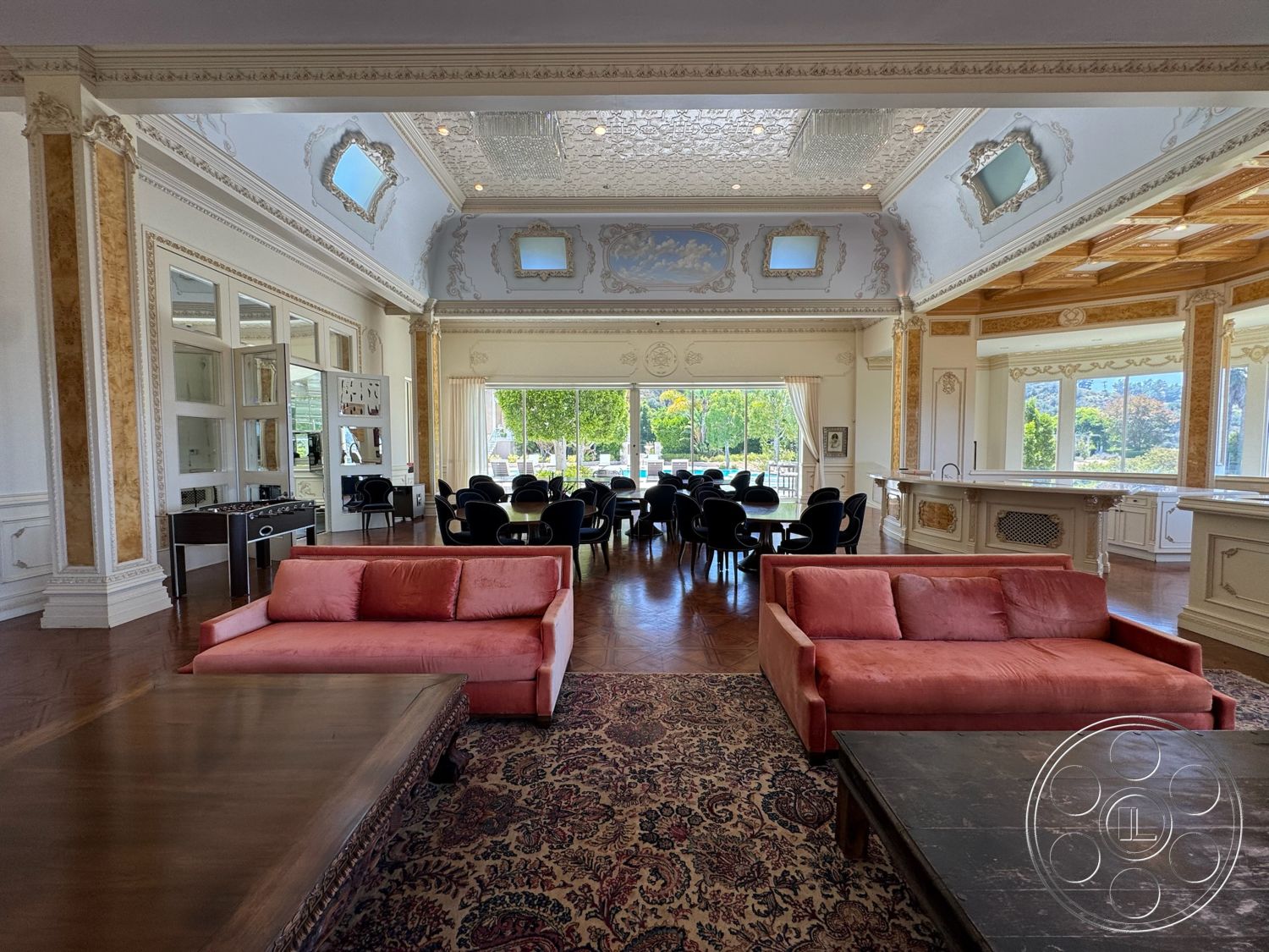 Beverly Compound - glass sliding doors, decorative wall art, chandelier lighting, dark wood coffee table, luxurious living space, open floor plan layout, traditional area rug, hardwood floor with dark finish, natural light-filled area, intricate wall moldings, ornate ceiling with decorative molding, wood-paneled ceiling, custom wood bar with cabinetry, large windows with outdoor views, pink upholstered sofa, round dining tables with black chairs, gold accents and details