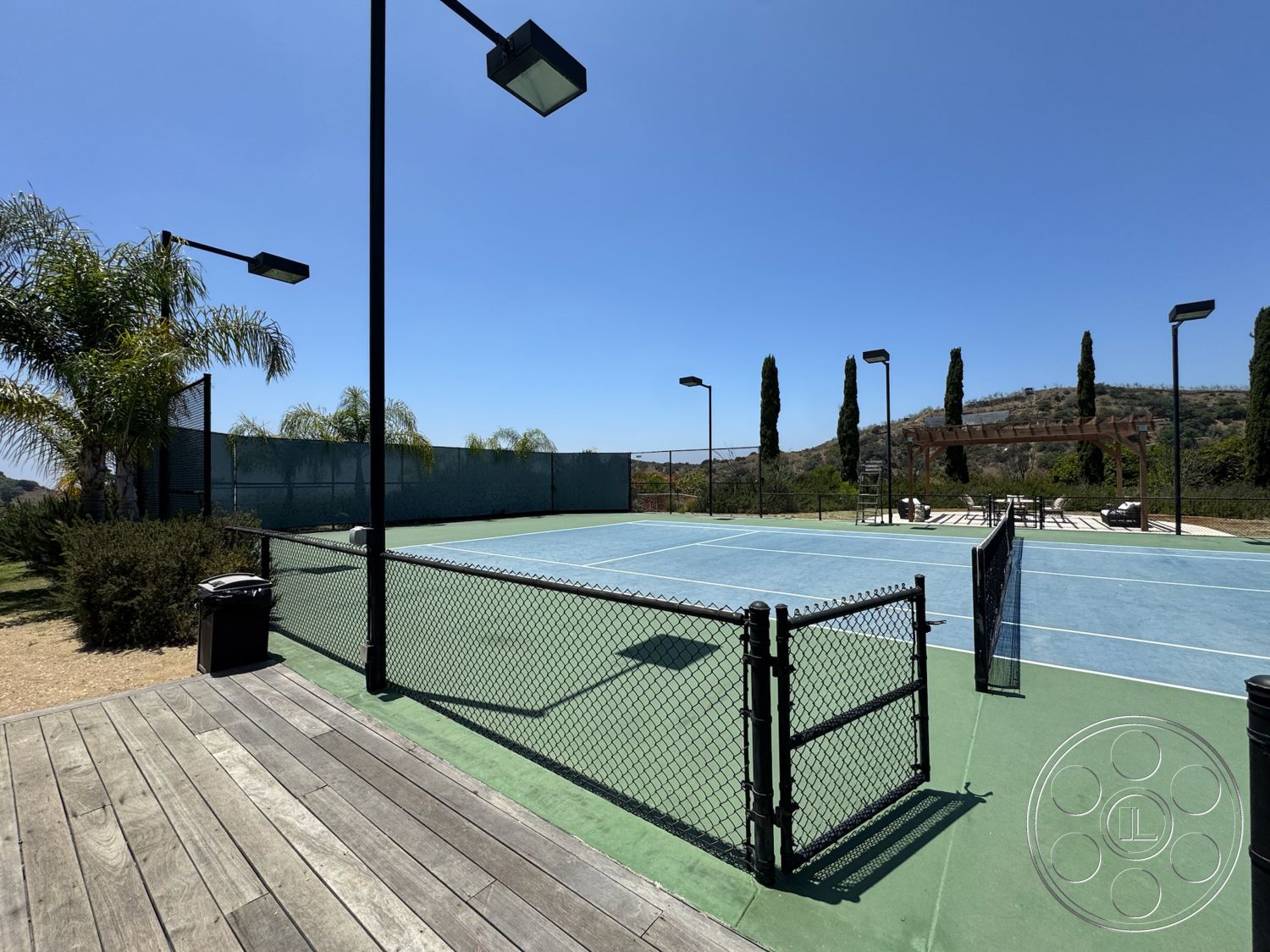 Beverly Compound - exterior, palm trees, outdoor lighting fixtures, chain link fence, mountain view setting, wooden deck flooring, landscape shrubs, pergola structure, tennis court surface, synthetic grass tennis court, open sky backdrop