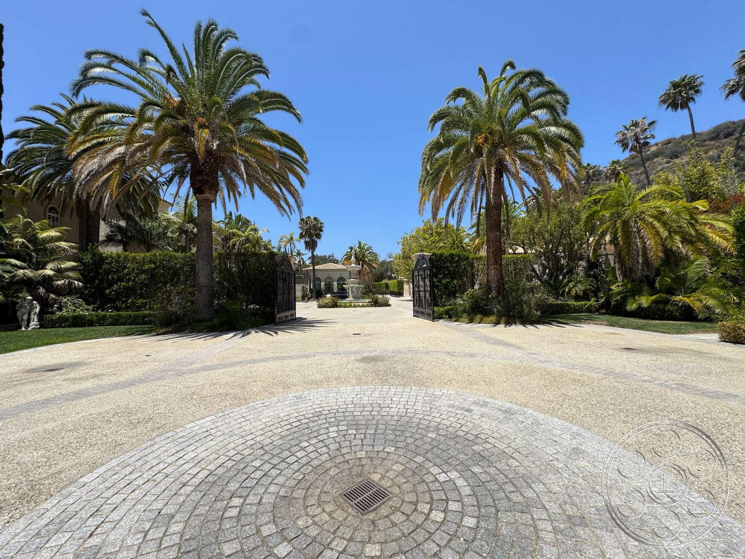 Beverly Compound - exterior, landscaped garden, lush greenery, palm trees, gravel pathways, gated entrance, decorative stone wall, cobblestone driveway, sunny outdoor setting, cobblestone circular patio