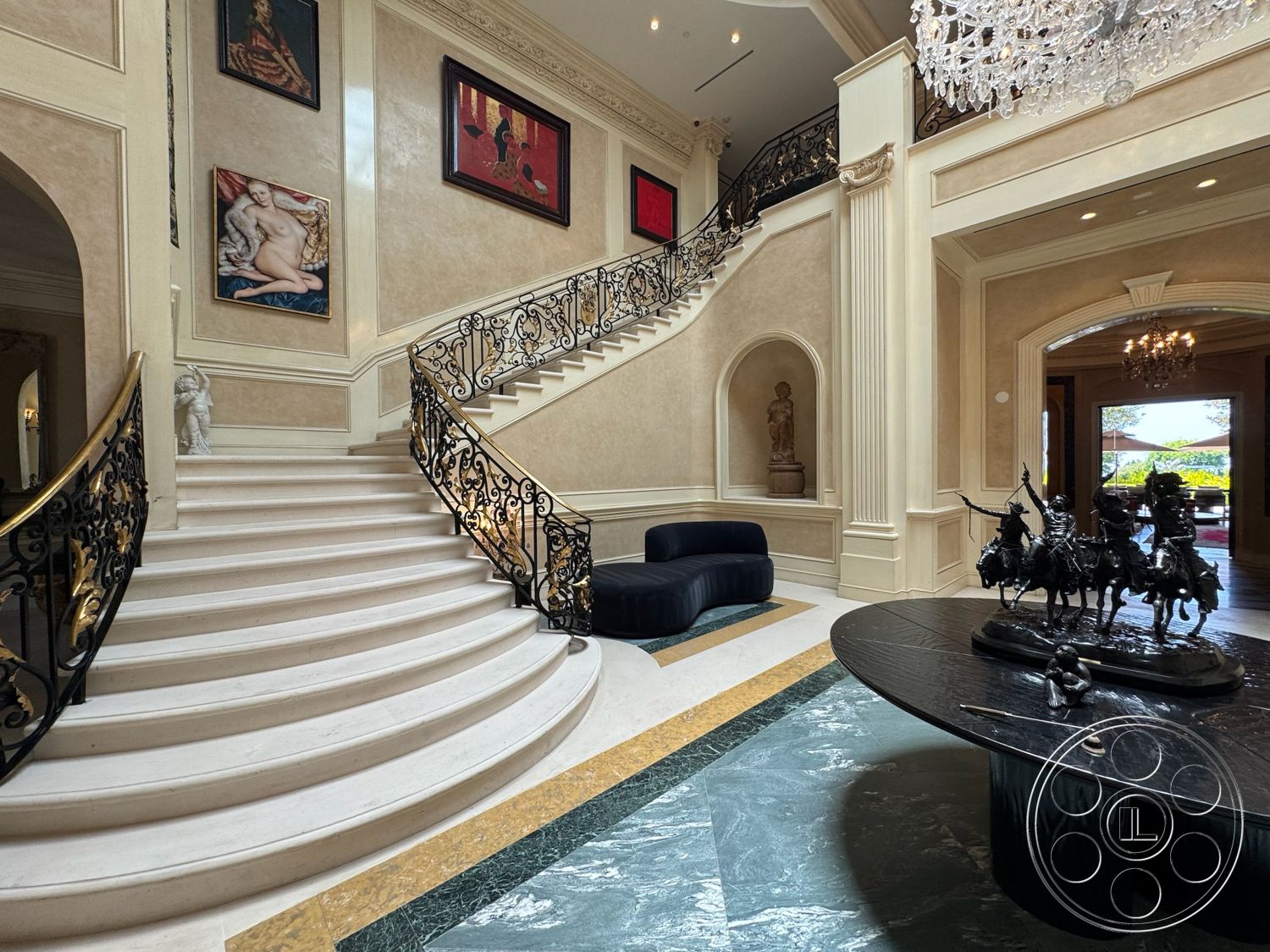 Beverly Compound - vaulted ceiling, chandelier lighting, artistic sculptures, textured plaster wall finish, decorative wall paintings, custom wrought iron staircase railing, curved marble staircase, stone floor entryway, black marble tabletop, gold accent border on flooring, sculptural wall niches, large glass door opening to outdoor area
