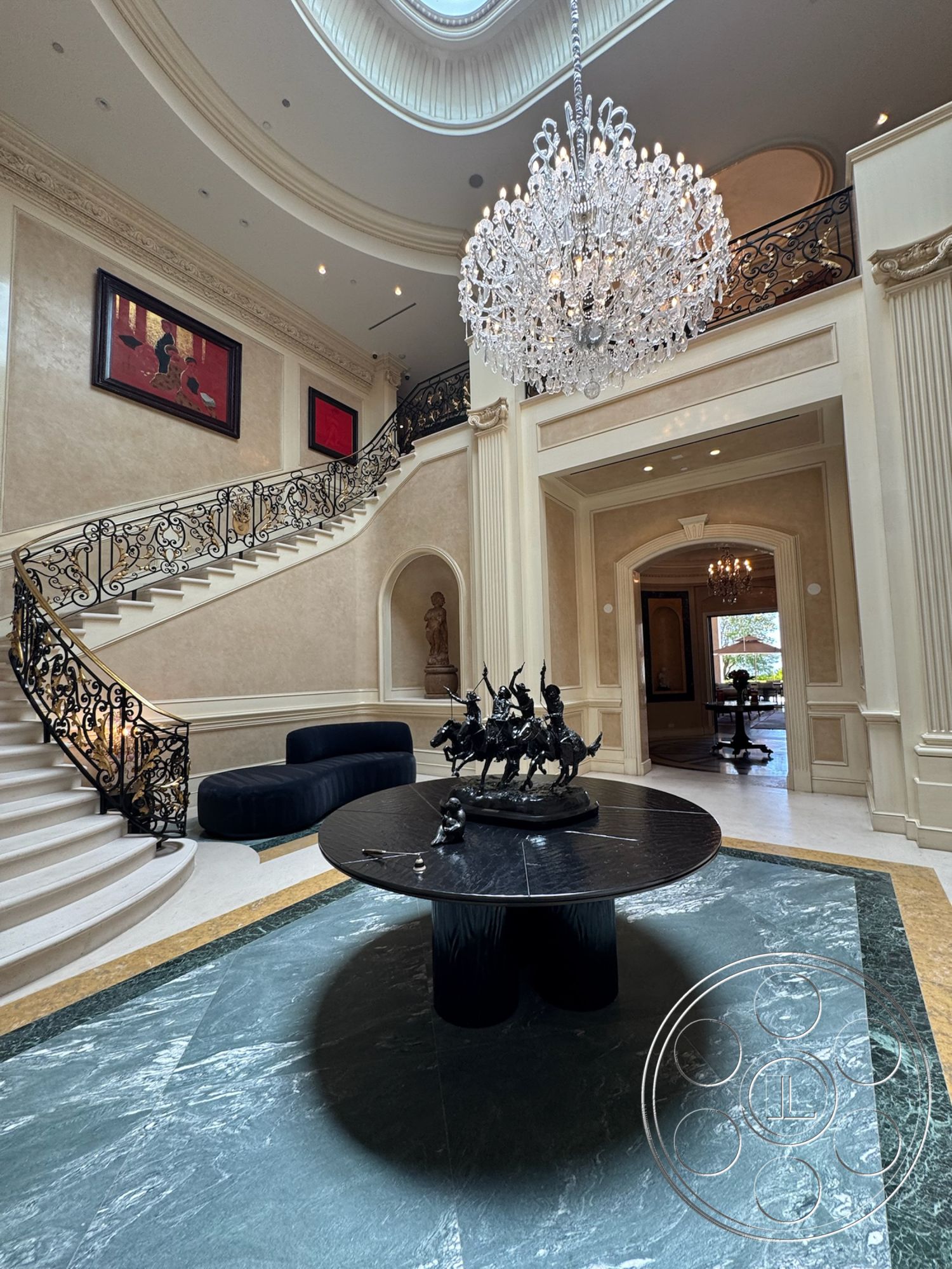 Beverly Compound - high ceilings, chandelier lighting, arched doorways, textured plaster walls, black leather sofa, curved staircase, cream-colored wall panels, statue decor, black marble round table, open foyer area, oval skylight ceiling, dark wrought iron railing, green marble floor, gold trim accents