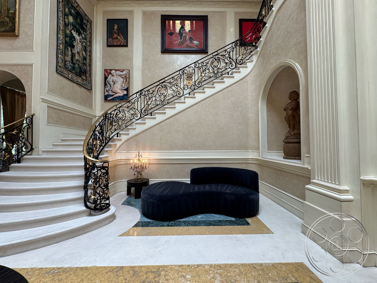 Beverly Compound - polished marble flooring, textured plaster walls, elegant chandelier lighting, intricate crown molding details, contemporary curved sofa, curved staircase with wrought iron railings, framed art pieces on walls, exposed wooden accents, decorative statuette, gold and blue decorative inlay, arched wall recess, large open foyer space