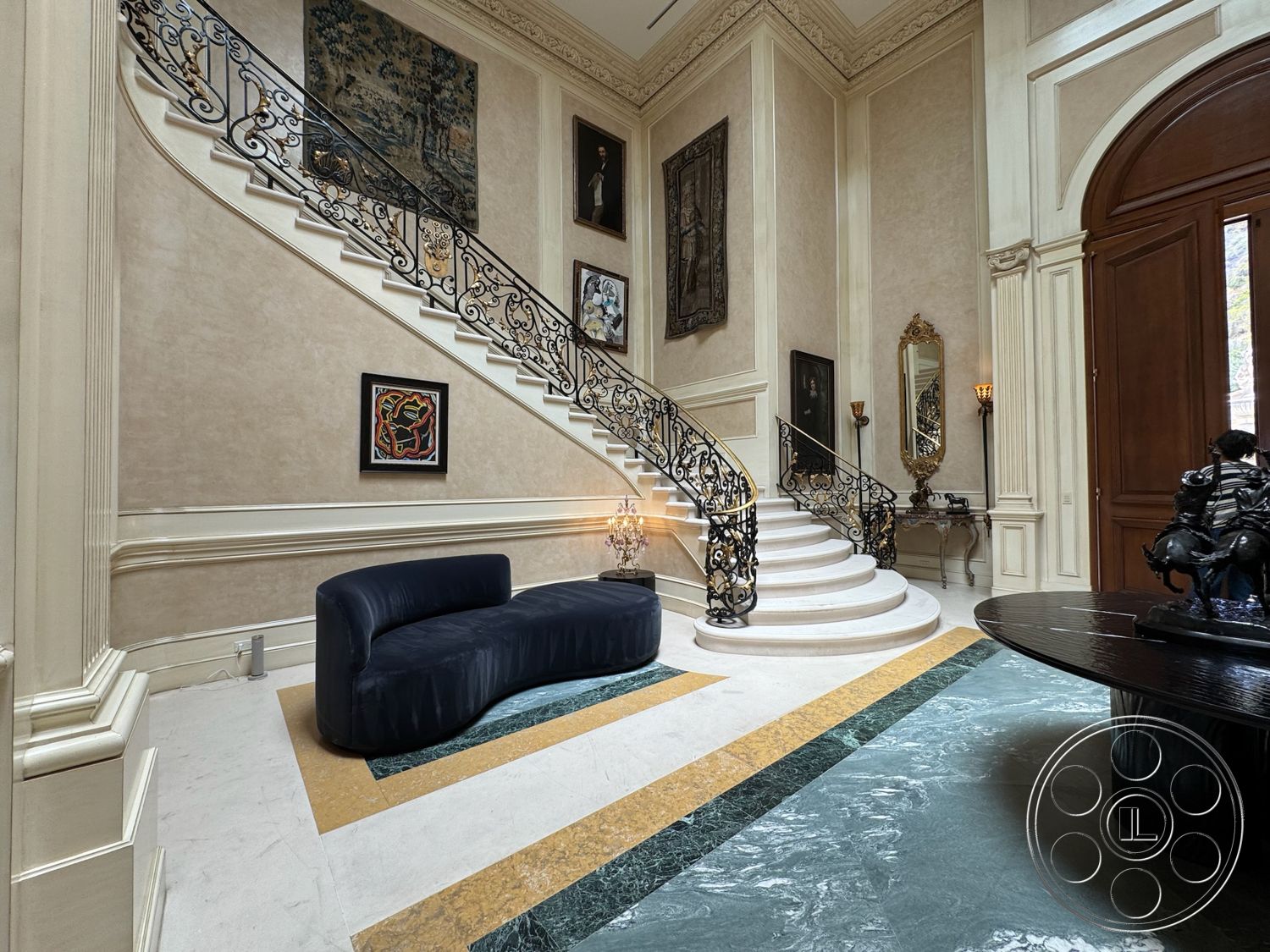 Beverly Compound - high ceilings, polished marble flooring, wooden double doors, dark wood console table, curved staircase with wrought iron railing, light-filled entryway, ornate crown molding details, cream-colored textured plaster walls, framed artworks on walls, sculptural decor piece, elegant candelabra sconces, geometric multi-colored marble inlay, luxurious foyer design