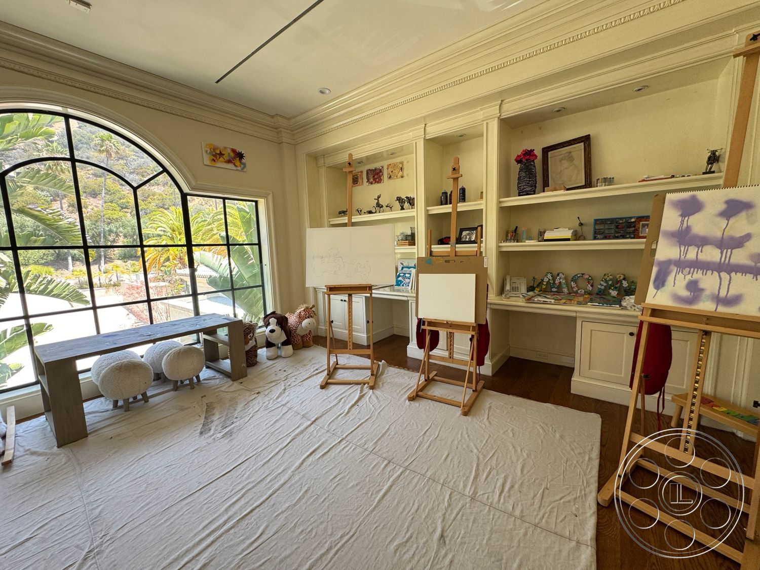 Beverly Compound - hardwood flooring, bright natural light, artist easels, built-in shelves, exposed wood ceiling beams, soft seating area, tropical garden view, arched window design, decorative art displays, painted wall panels, fabric drop cloth, wood table with industrial design, spacious artist studio
