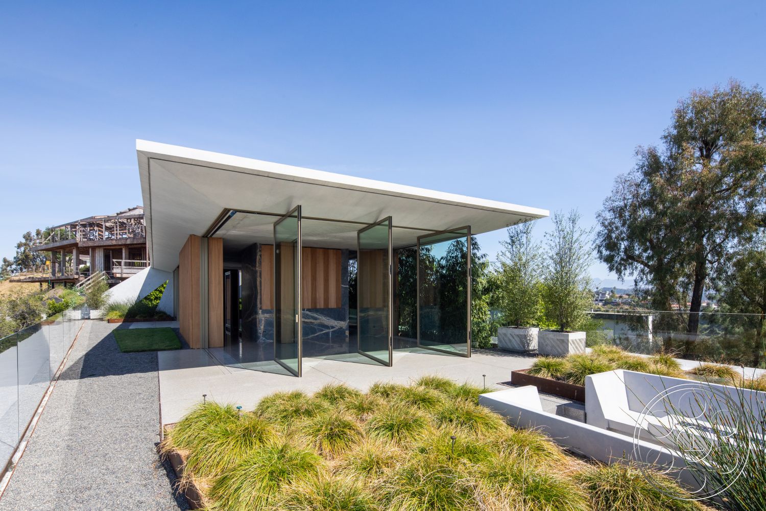 Modern 516 - exterior, flat roof, minimalist design, wood paneling, concrete flooring, large glass windows, surrounding trees, gravel pathway, modern architectural style, open view, hilltop location, outdoor planters, landscaped garden area, glass entrance doors, natural grass landscaping, rectangular concrete steps, step stone patio