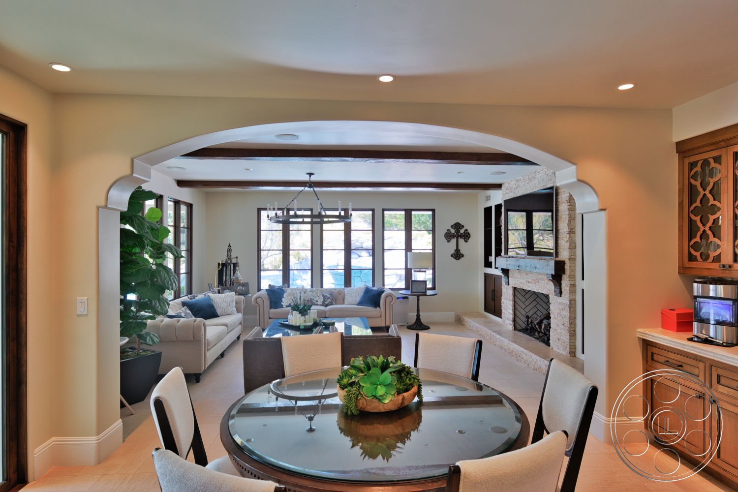 Hayden House - upholstered dining chairs, floor-to-ceiling windows, glass dining table, textured plaster walls, arched doorway, modern chandelier, vaulted ceiling with exposed wood beams, decorative plant accents, neutral tile flooring, stone fireplace feature wall, wood cabinetry in kitchen area