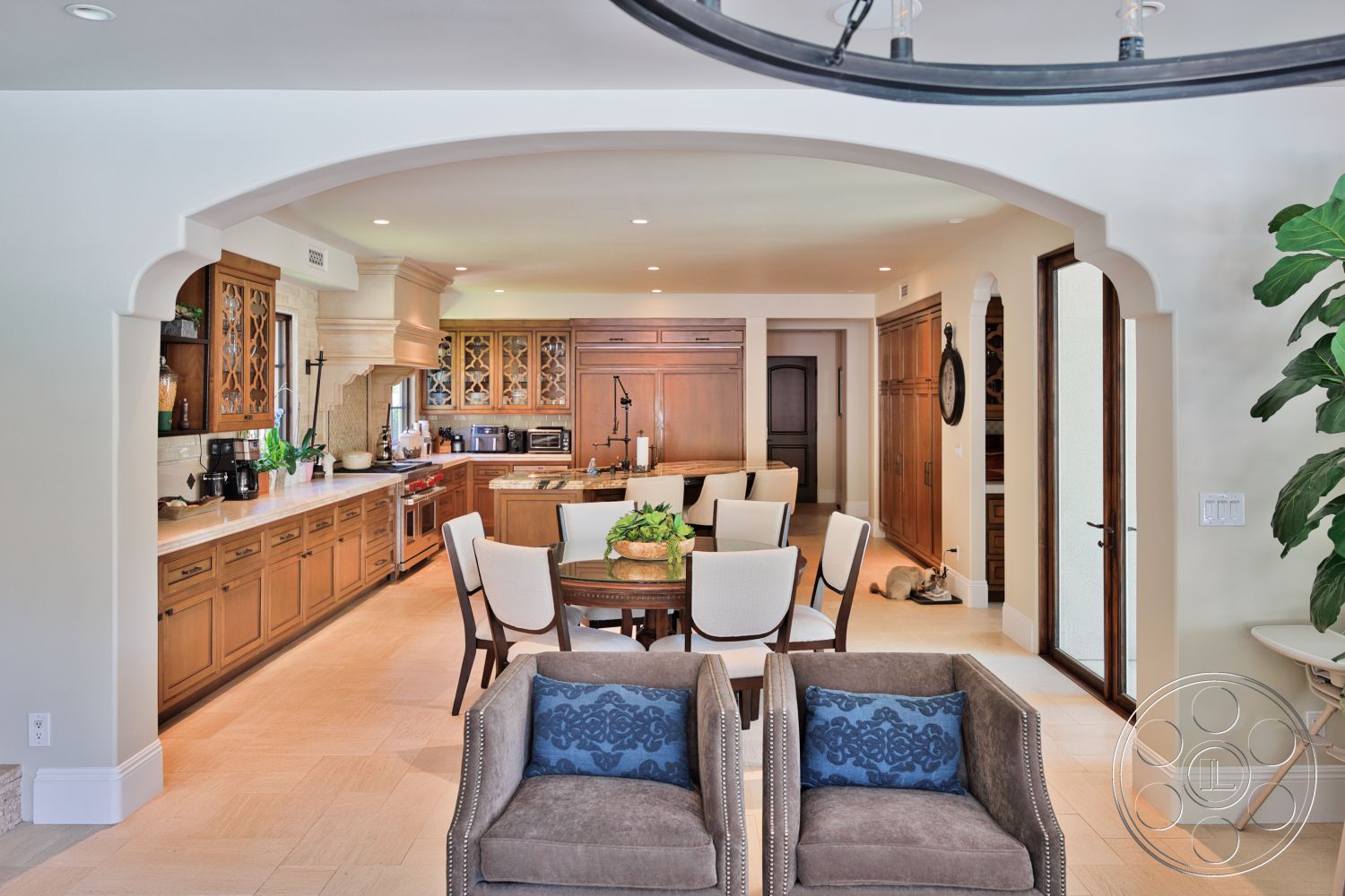 Hayden House - open floor plan, stainless steel appliances, upholstered dining chairs, round dining table, natural stone flooring, arched doorway, granite kitchen countertops, wooden door frames, potted indoor plant, kitchen with breakfast bar, cream-colored textured plaster walls, custom wood kitchen cabinets, decorative wood beam ceiling, living area with soft seating