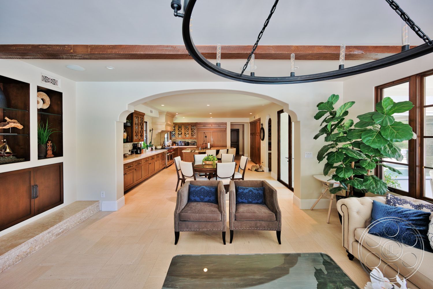 Hayden House - stone accent wall, large windows for natural light, decorative indoor plant, arched doorway, soft beige sofa, chandelier light fixture, hardwood living room floors, neutral painted walls, wood beam ceiling, built-in wood cabinetry, glass display shelves, open concept kitchen area, plush armchairs with blue accents