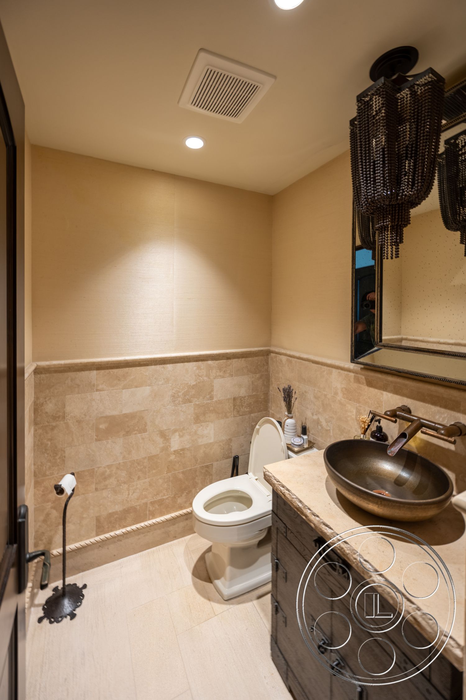 Hayden House - wooden flooring, textured plaster walls, white porcelain toilet, interior bathroom, modern chandelier lighting, light wood vanity cabinet, travertine wall tiles, stone countertop sink, round bronze vessel sink, framed mirror with ornate detailing