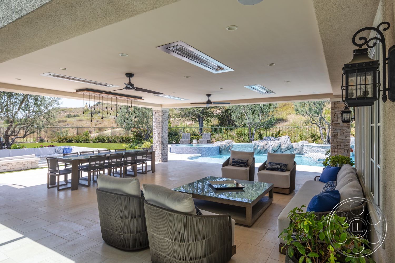 Hayden House - exterior, white stucco walls, wood dining table, al fresco dining area, woven outdoor chairs, landscaped garden area, concrete patio flooring, open patio space, exposed ceiling lights, glass patio table, lounge seating area, natural stone wall accents, stone column details, stainless steel outdoor ceiling fans, swimming pool with rock features
