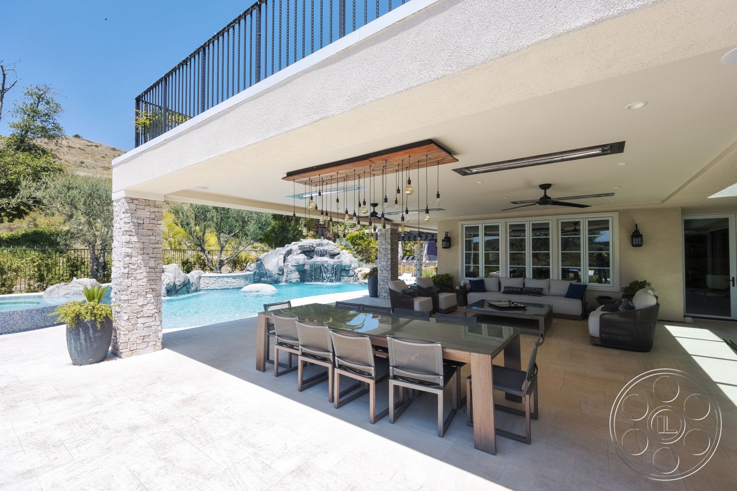 Hayden House - exterior, glass sliding doors, outdoor seating area, rectangular dining table, decorative potted plants, concrete patio flooring, landscaped garden setting, metal railing balcony, mountain view backdrop, plastered wall finish, hanging pendant lighting, stone column supports, contemporary outdoor kitchen area, swimming pool with rock waterfall feature