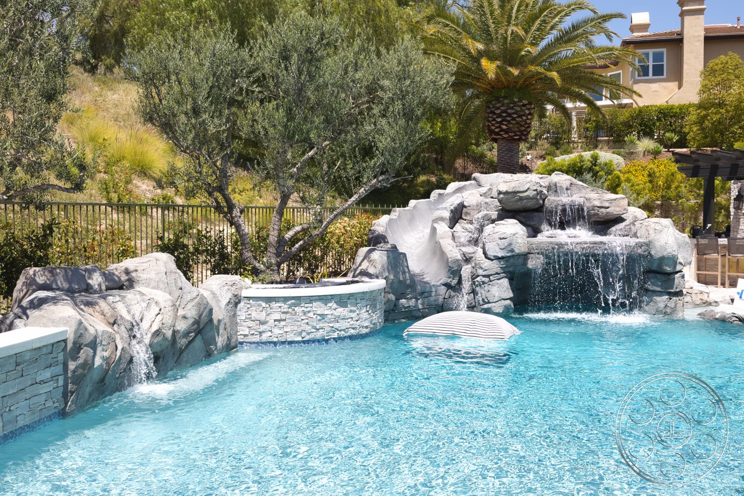 Hayden House - exterior, sunny outdoor setting, concrete pool border, lush landscaping with palm trees, metal fence enclosure, tile pool decking, stone waterfall feature, natural stone rock formations, fiberglass pool
