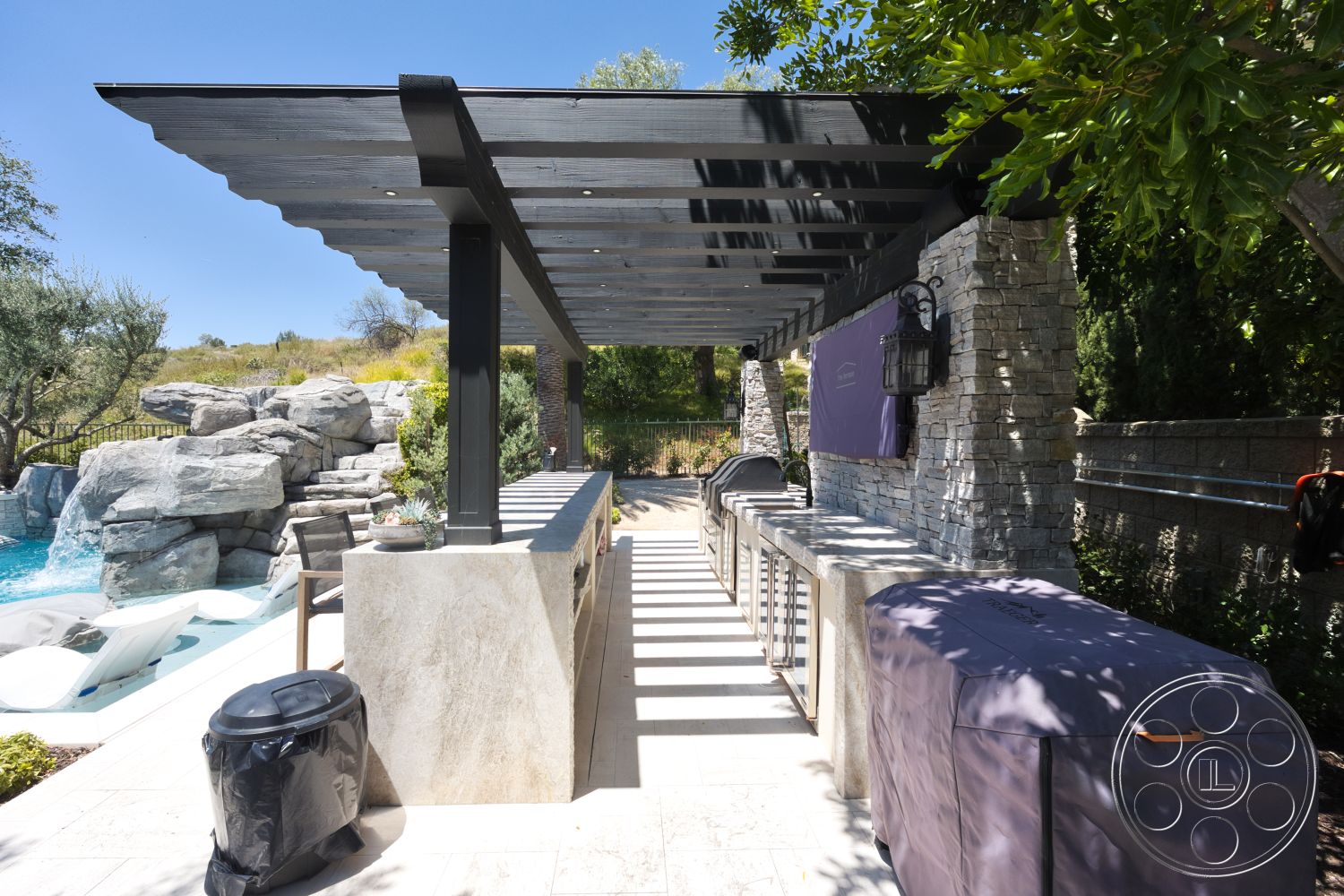 Hayden House - exterior, lush greenery backdrop, landscaped garden setting, stone patio flooring, modern outdoor dining space, outdoor grill area, natural rock formation, stone pillar supports, horizontal wood trellis roof, outdoor kitchen countertop, concrete outdoor bar, vintage outdoor lantern, pool area with stone features