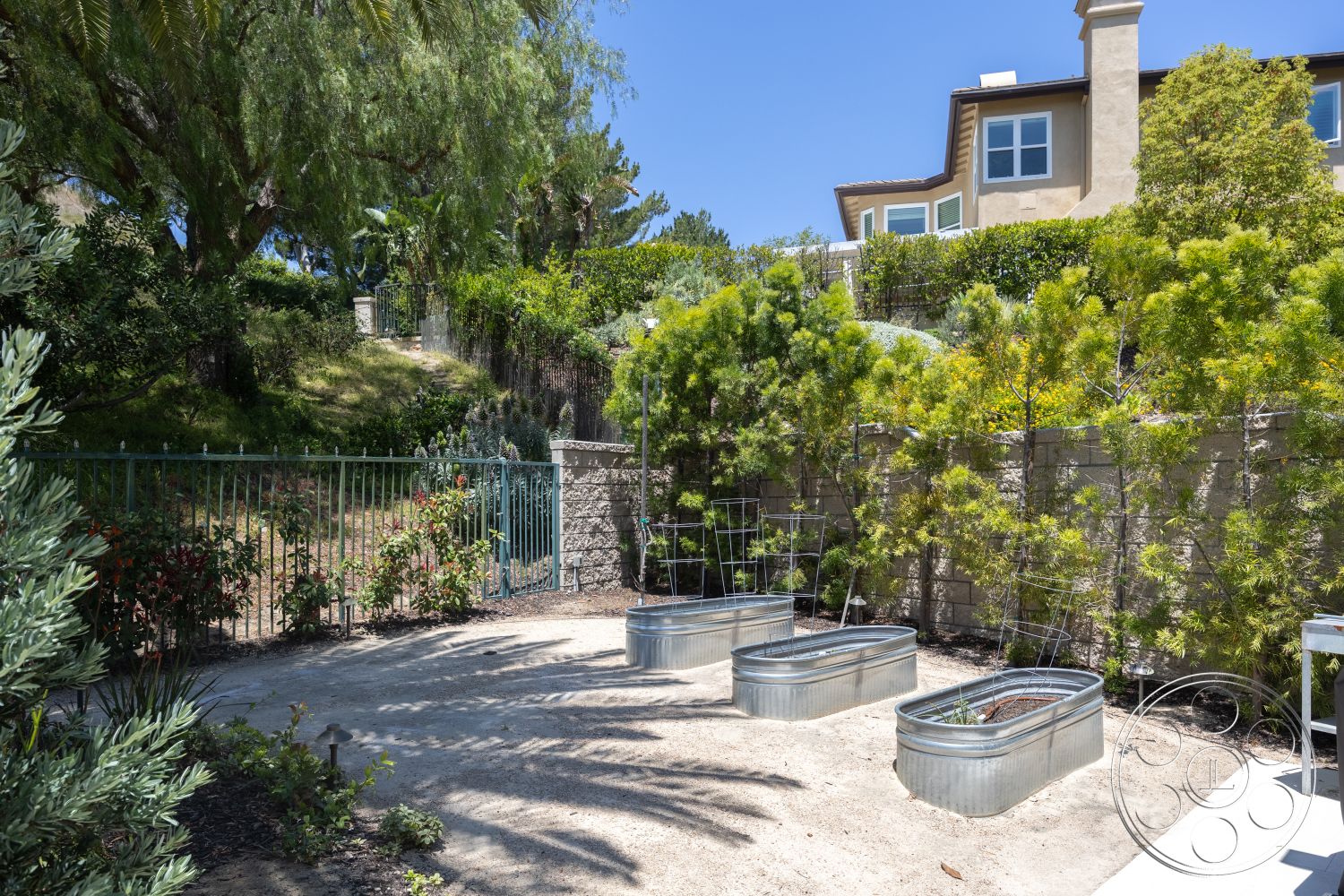 Hayden House - exterior, sloped terrain, mature trees, garden setting, natural sunlight, paved outdoor area, circular stone planters, lush green landscaping, concrete retaining wall, stucco house facade, galvanized metal raised garden beds, sandblasted gravel path, iron garden gate, decorative trellises