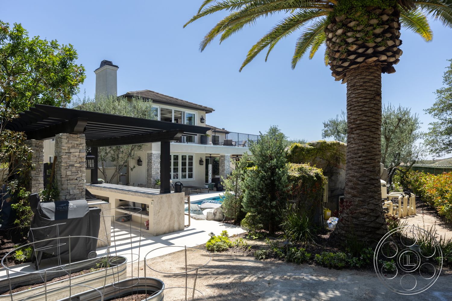 Hayden House - exterior, rectangular swimming pool, concrete patio flooring, natural stone wall accents, palm trees in landscaping, stone outdoor kitchen countertop, open backyard setting, exposed stone column supports, black wooden pergola structure, two-story Mediterranean style home, multi-paned French windows, modern outdoor grill station, surrounding garden flower beds