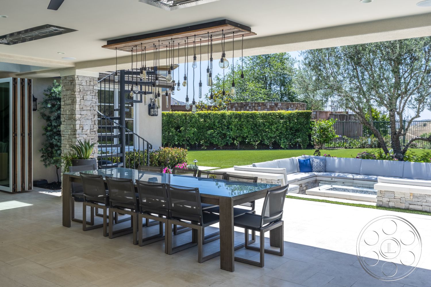 Hayden House - exterior, stone accent wall, glass dining table, lawn area, lush greenery backdrop, modern outdoor seating, curved landscaping, wood dining chairs, concrete patio flooring, landscaped garden beds, open patio area, decorative outdoor planters, hanging pendant light fixtures, framed glass doors, suspended ceiling with lights, stainless steel outdoor stair railings, freestanding hot tub