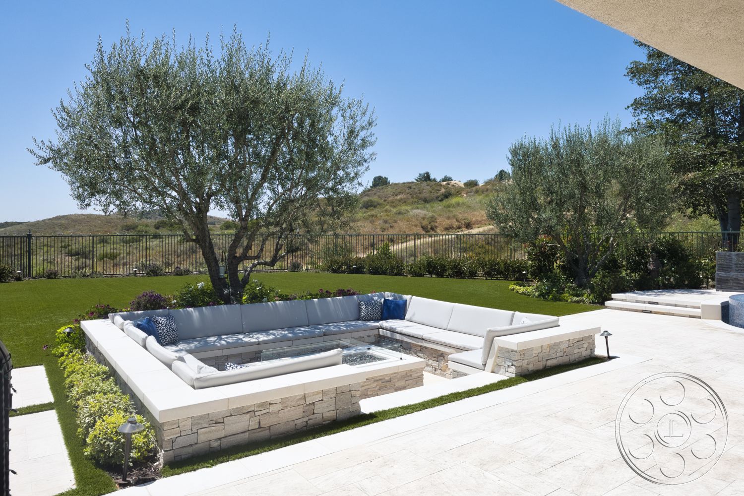 Hayden House - exterior, outdoor seating area, mountain view, olive trees, decorative potted plants, concrete patio flooring, fenced backyard, landscaped garden beds, sunny hillside location, rectangular fire pit, artificial grass lawn, decorative outdoor cushions, stone fire pit seating area, light-colored stone retaining wall