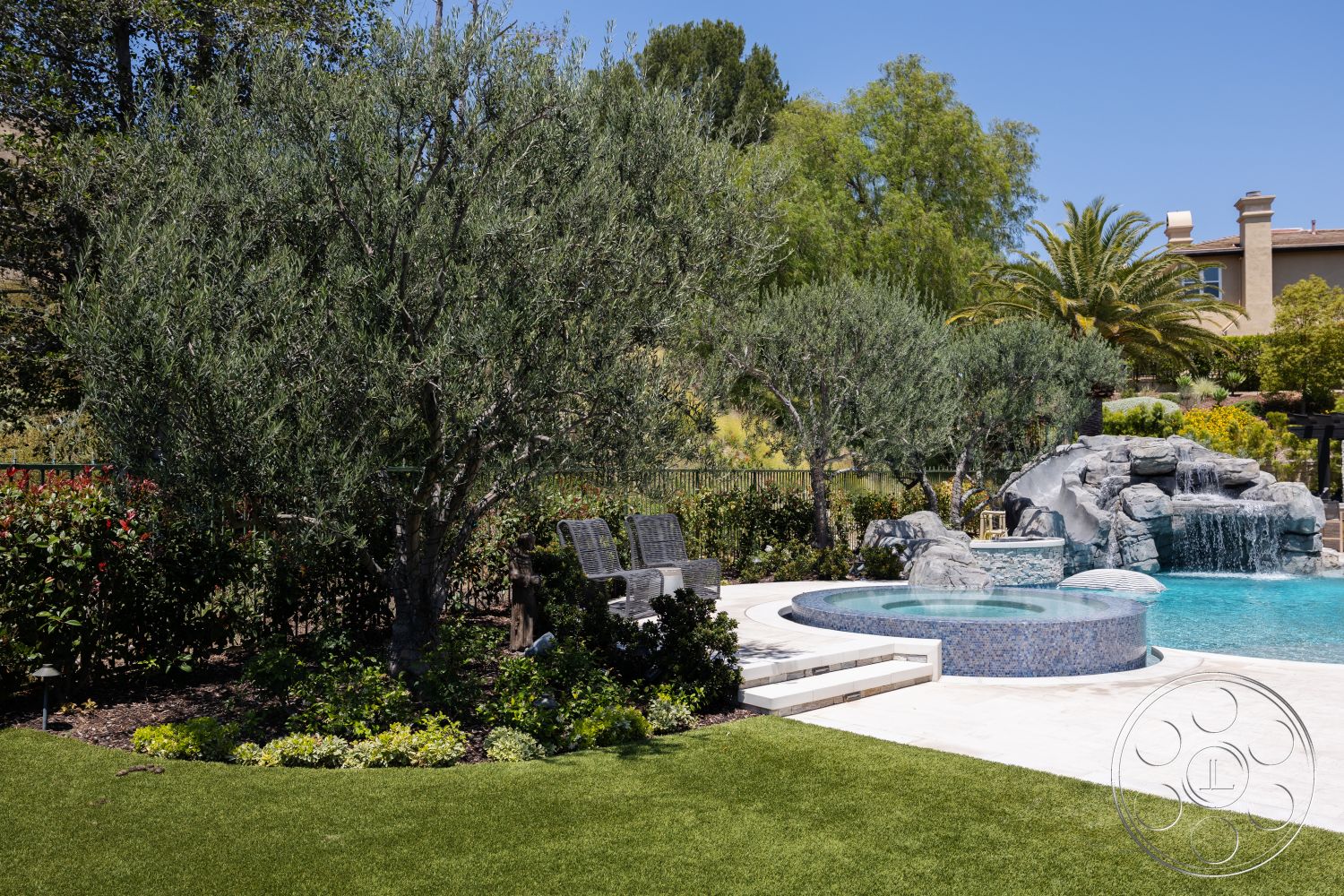 Hayden House - exterior, wooden lounge chairs, rectangle swimming pool, artificial grass lawn, concrete patio surround, stone waterfall feature, landscaped garden with flowering shrubs, round spa with mosaic tile finish, fenced backyard setting, variety of trees including olive trees