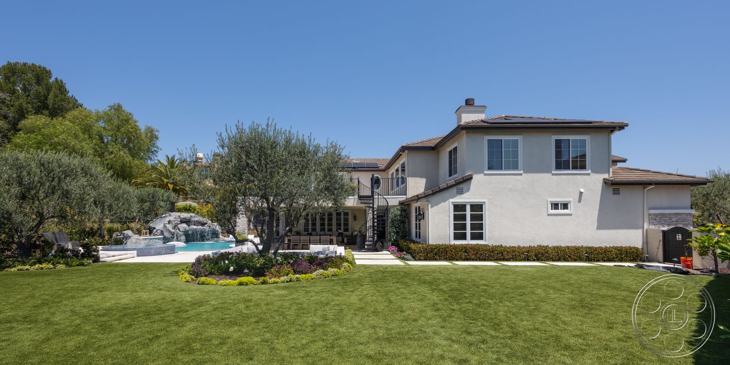Hayden House - exterior, landscaped garden, outdoor seating area, rectangular swimming pool, lush green lawn, tropical landscaping, stucco walls, arched doorways, tile roof, hardscaped pathway, paved pool deck, two-story home, fenced backyard, decorative stone accents, natural stone water feature, urban outdoor setting, plantation shutters