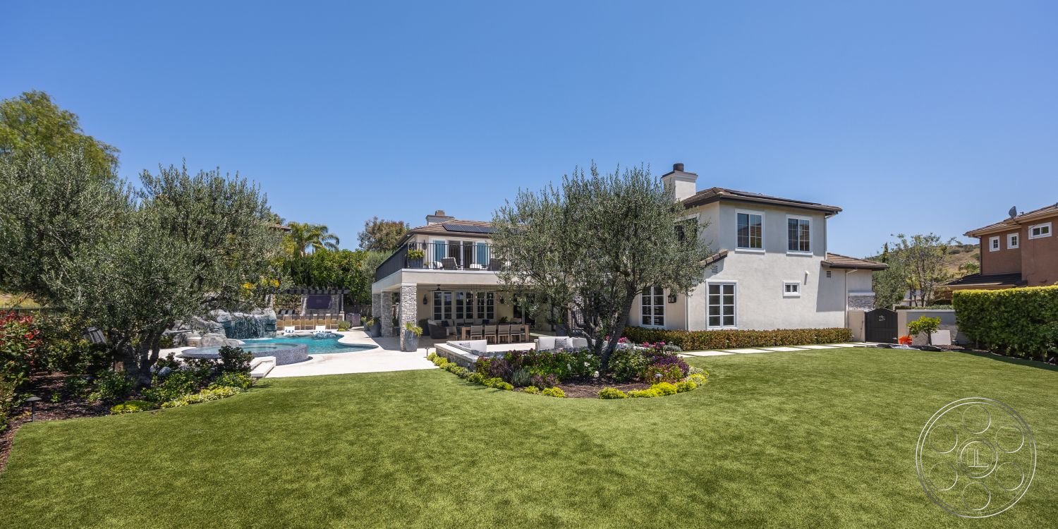 Hayden House - large windows, exterior, outdoor seating area, rectangular swimming pool, sloped roof, modern architectural style, suburban backyard, stone pool deck, landscaped garden beds, paved patio area, mature olive trees, green hedge border, synthetic grass lawn, wood patio furniture, stucco house walls