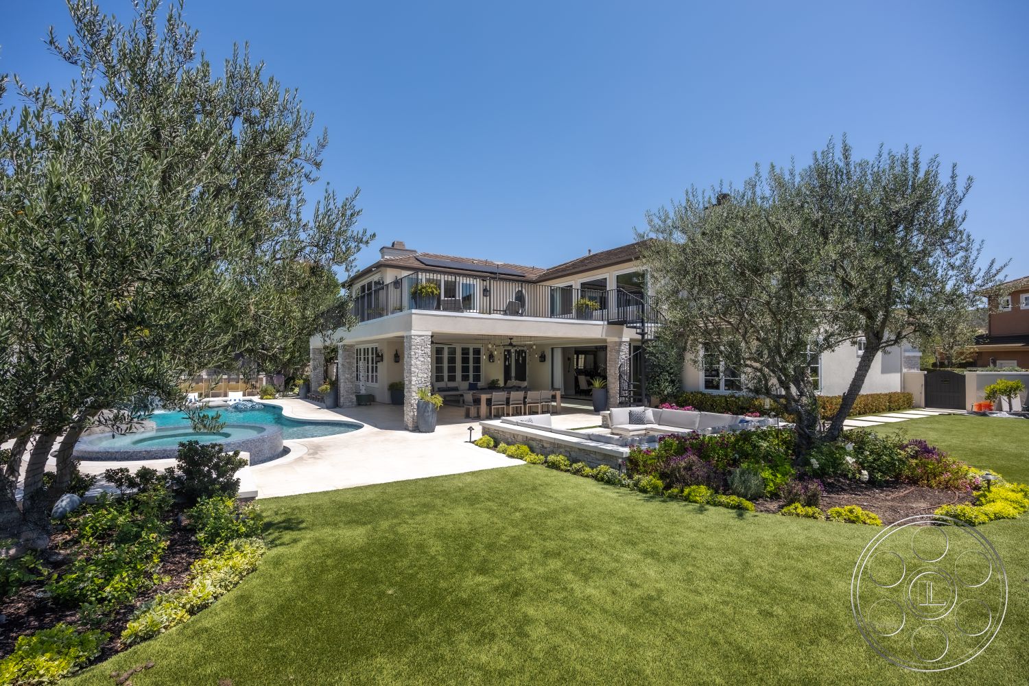 Hayden House - exterior, glass sliding doors, outdoor seating area, covered porch, landscaped backyard, stucco walls, concrete pool deck, artificial turf lawn, two-story home, oval swimming pool, flower garden beds, metal railing balcony, paved patio flooring, stone accent pillars