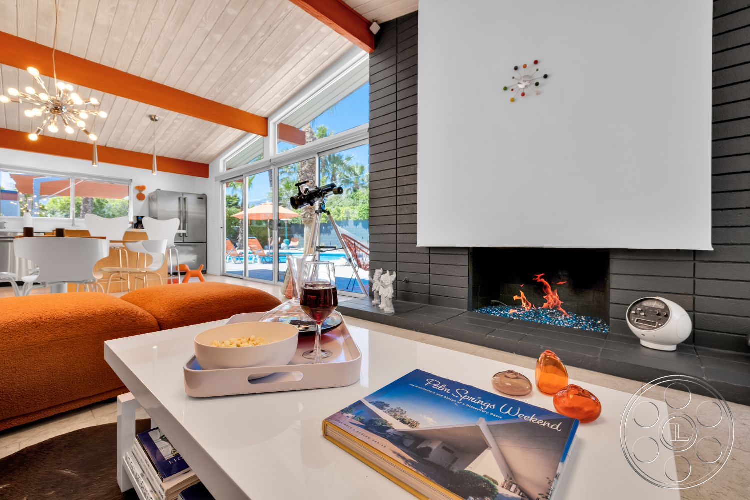 Palm Springs 76 - concrete flooring, modern chandelier, swimming pool view, orange fabric sofa, glass slider doors, white coffee table, exposed wood ceiling beams, vaulted living room ceiling, open floor plan living area, glass decorative objects, black brick fireplace wall