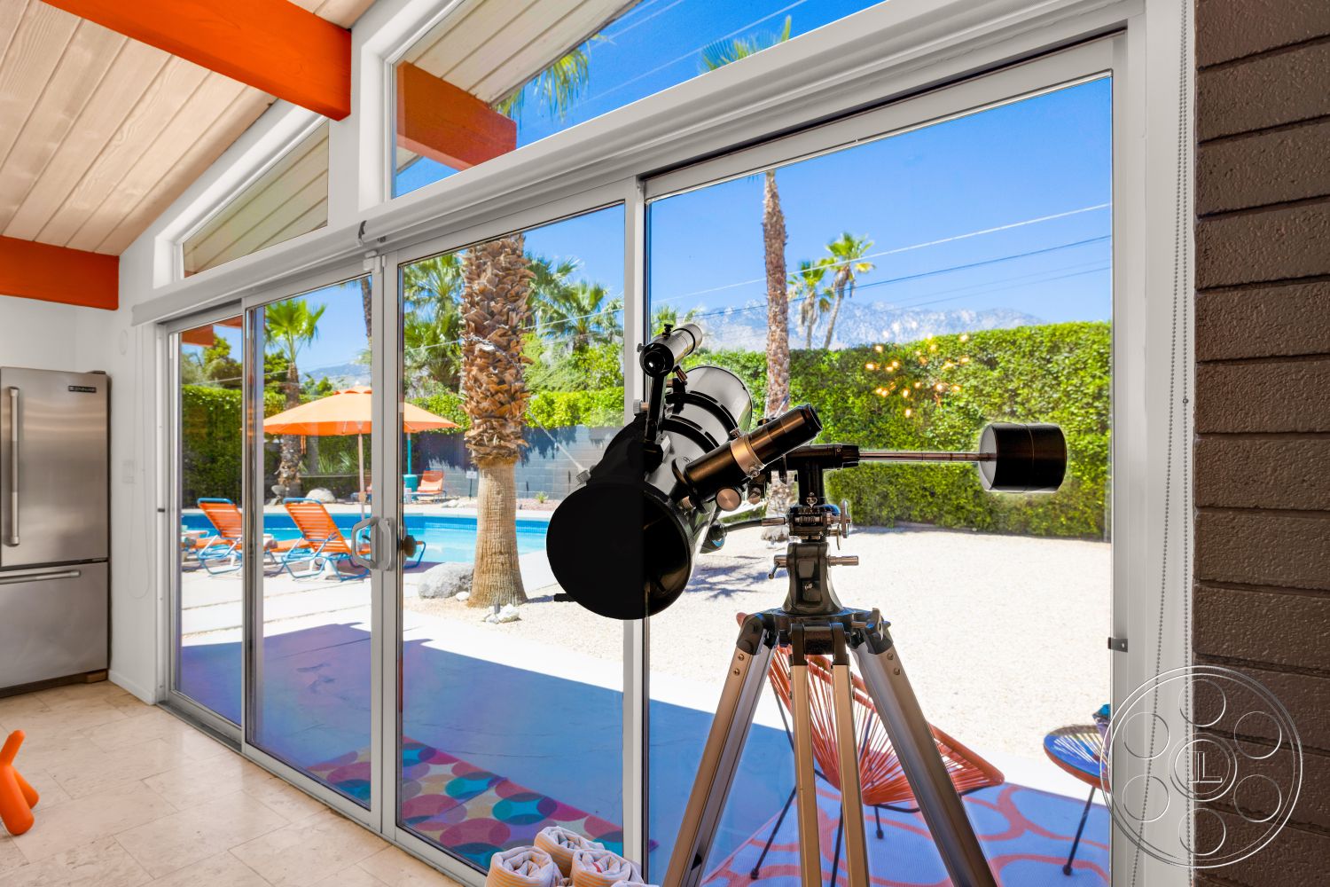 Palm Springs 76 - exterior, landscaped garden, sliding glass doors, rectangular swimming pool, tropical palm trees, outdoor lounge chairs, stone pool deck, white painted walls, concrete patio floor, decorative patterned rug, vaulted ceiling with wood beam details, metal telescope on tripod stand, orange patio umbrellas, mountain view in the background, bright daylight setting