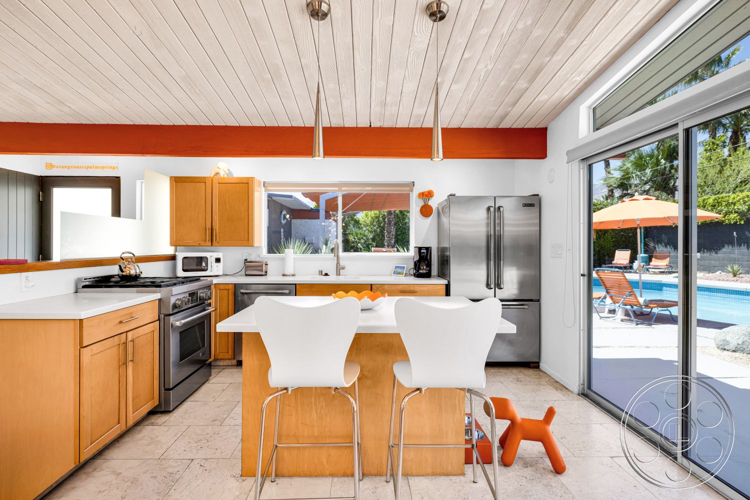 Palm Springs 76 - modern kitchen, natural light, landscaped garden, open floor plan, stainless steel appliances, wood cabinetry, tropical setting, white quartz countertop, poolside view, stone tile flooring, floor-to-ceiling sliding glass doors, outdoor patio area, vaulted ceiling with wood beams, orange accent beam, breakfast bar with white stools