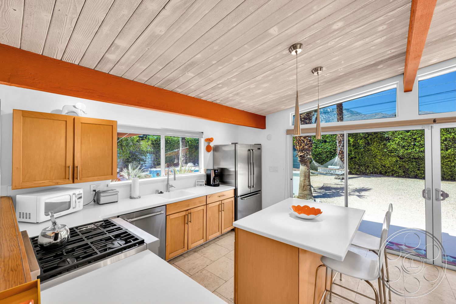 Palm Springs 76 - large windows, stainless steel appliances, open concept layout, modern pendant lighting, light wood cabinets, beige tile flooring, palm tree view, interior kitchen, white quartz countertops, exposed wooden ceiling beams, white bar stools, orange accent beams