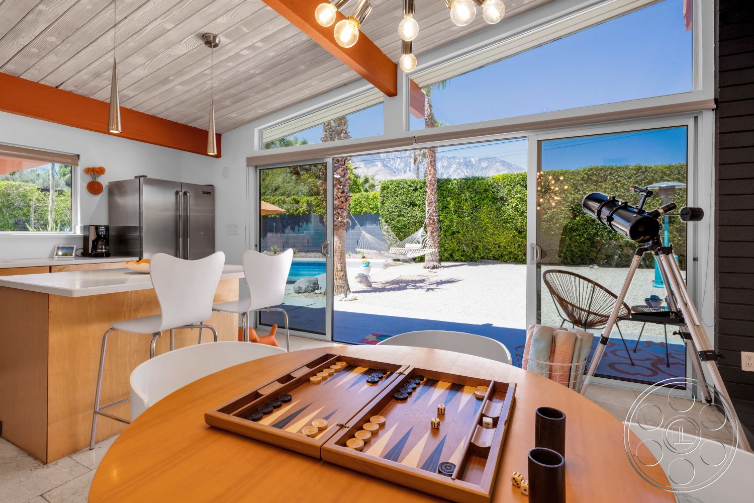 Palm Springs 76 - modern kitchen, large windows for natural light, ceramic tile flooring, stainless steel refrigerator, glass sliding door, round wooden dining table, outdoor pool area, gravel patio flooring, light wood kitchen cabinetry, white quartz kitchen countertop, white plastic dining chairs, palm trees in outdoor setting, wood ceiling with exposed beams, mountain view from windows, hammock visible in outdoor space