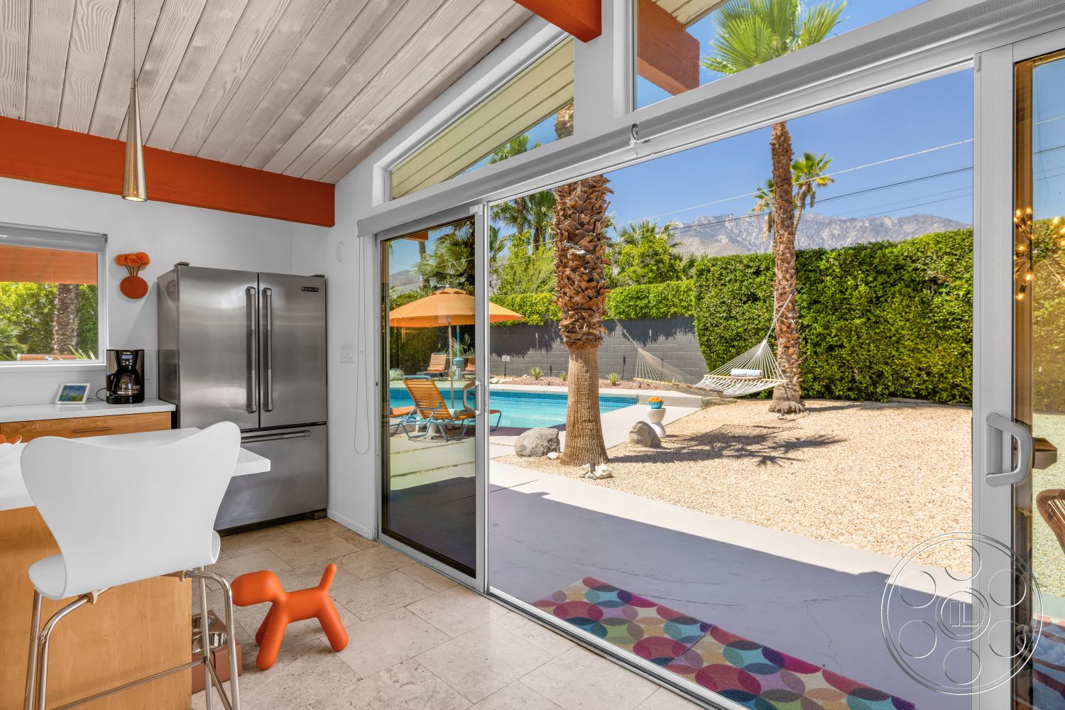 Palm Springs 76 - concrete floor, glass sliding doors, rectangular swimming pool, modern architectural style, stainless steel refrigerator, colorful area rug, mountain view setting, wood beam ceiling, kitchen countertop, white modern dining chair, planted palm trees visible, surrounding gravel patio