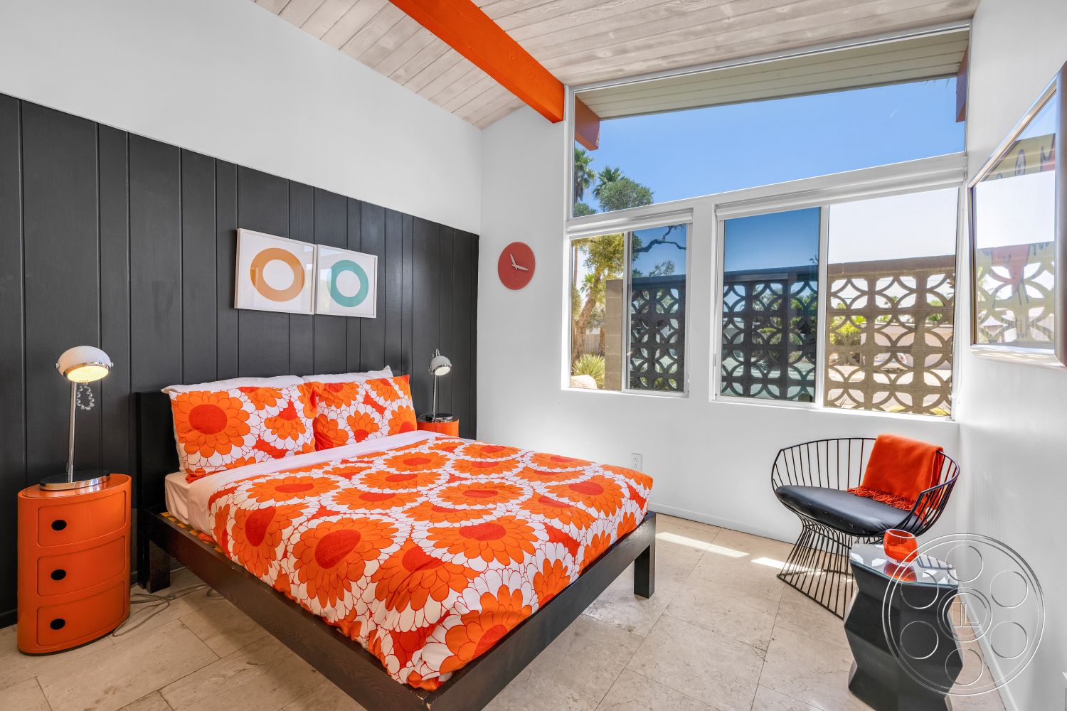 Palm Springs 76 - neutral walls, bright color accents, tile bedroom flooring, metal accent chair, contemporary bedside lamps, black wood paneling accent wall, high vaulted ceiling with exposed wood beams, modern bedding with floral pattern, large windows with geometric window pattern, minimalist orange circular wall clock, orange nightstand