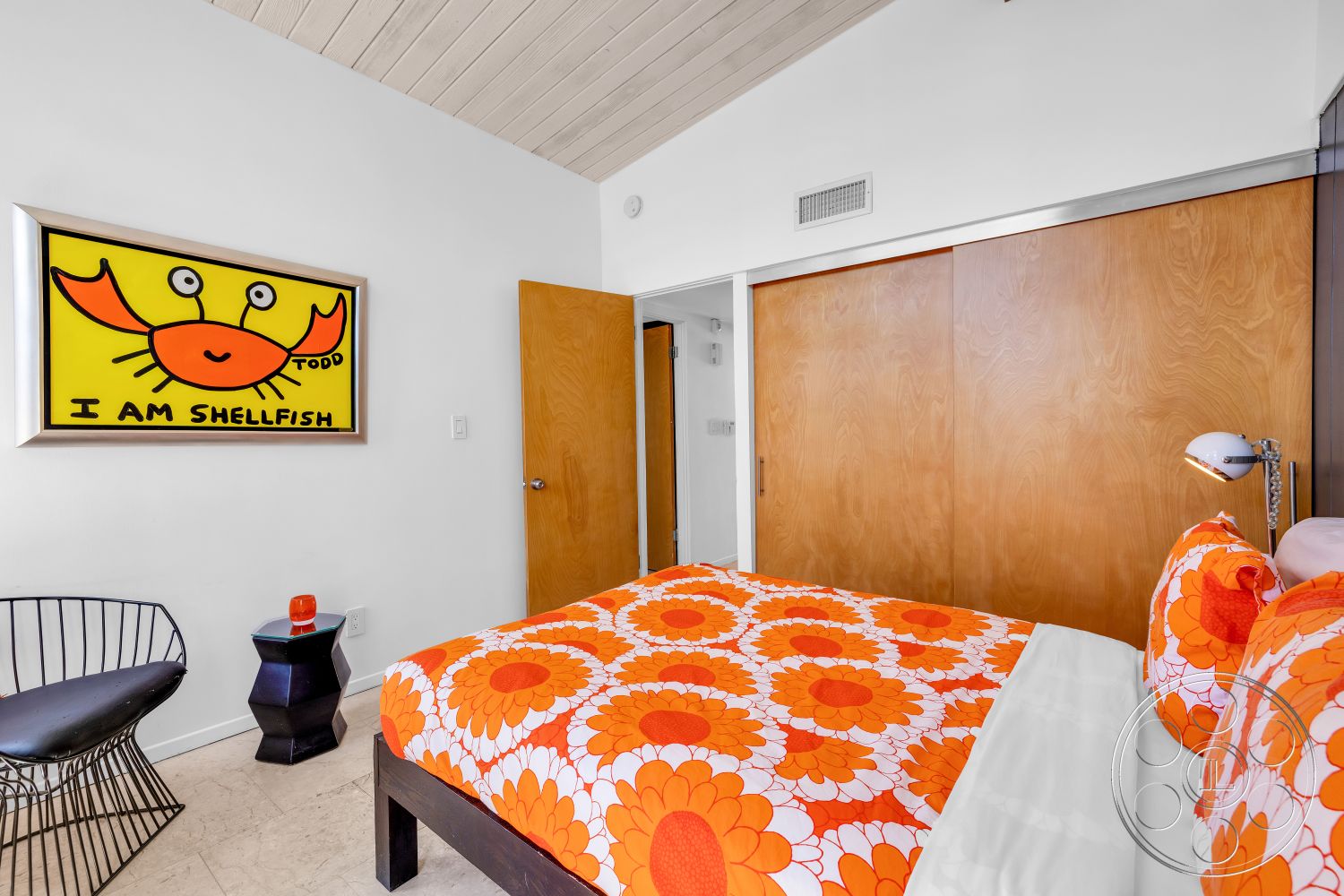 Palm Springs 76 - hardwood bedroom floors, white painted walls, vaulted ceiling with wood paneling, wood sliding closet doors, colorful artwork on wall, metal wire chair, black side table, orange floral bedding