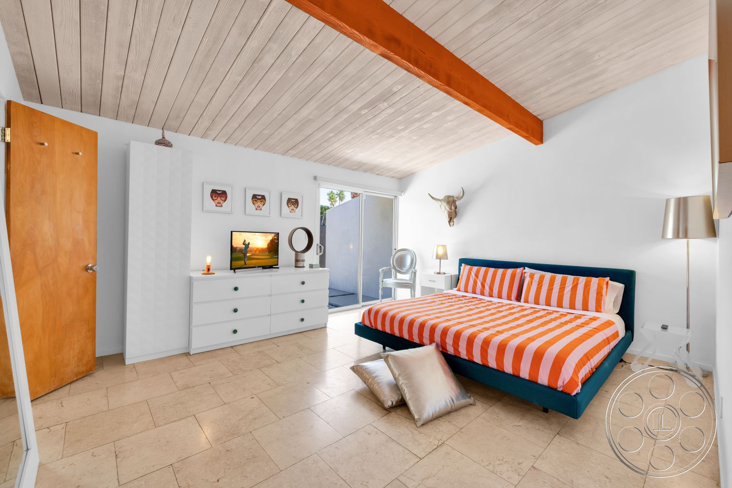 Palm Springs 76 - clear acrylic chair, wooden door, light wood dresser, gray accent pillows, exposed wood ceiling beams, white painted walls, framed artwork on wall, tile bedroom flooring, modern bedside lamp, blue upholstered bed frame, orange striped bedding, floor-to-ceiling sliding glass door, decorative wall mounted bull skull