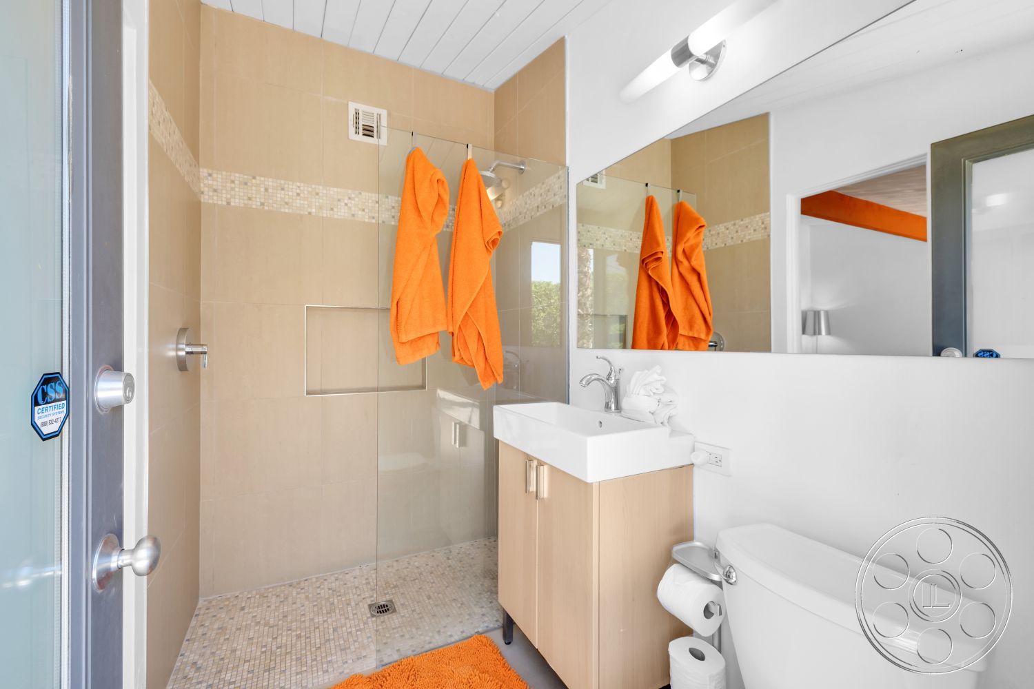 Palm Springs 76 - light wood cabinetry, white toilet, modern light fixture, interior bathroom, white painted walls, mirror above sink, transparent glass shower enclosure, tile shower walls, natural stone floor tiling, modern vanity with white countertop, orange towels hanging, orange bath mat, sloped ceiling with wood paneling