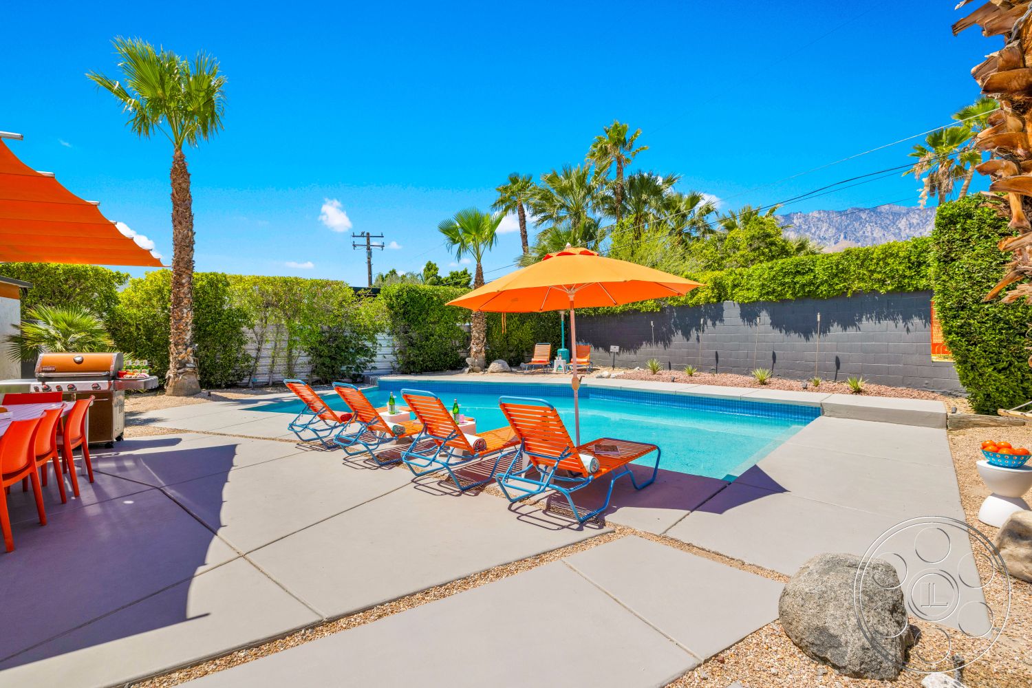 Palm Springs 76 - exterior, palm trees, barbecue grill, gray block wall, gravel landscaping, rectangular pool, concrete pool border, concrete patio floor, orange outdoor lounge chairs, orange pool umbrellas, low shrubbery, colorful outdoor dining set, sunny desert setting