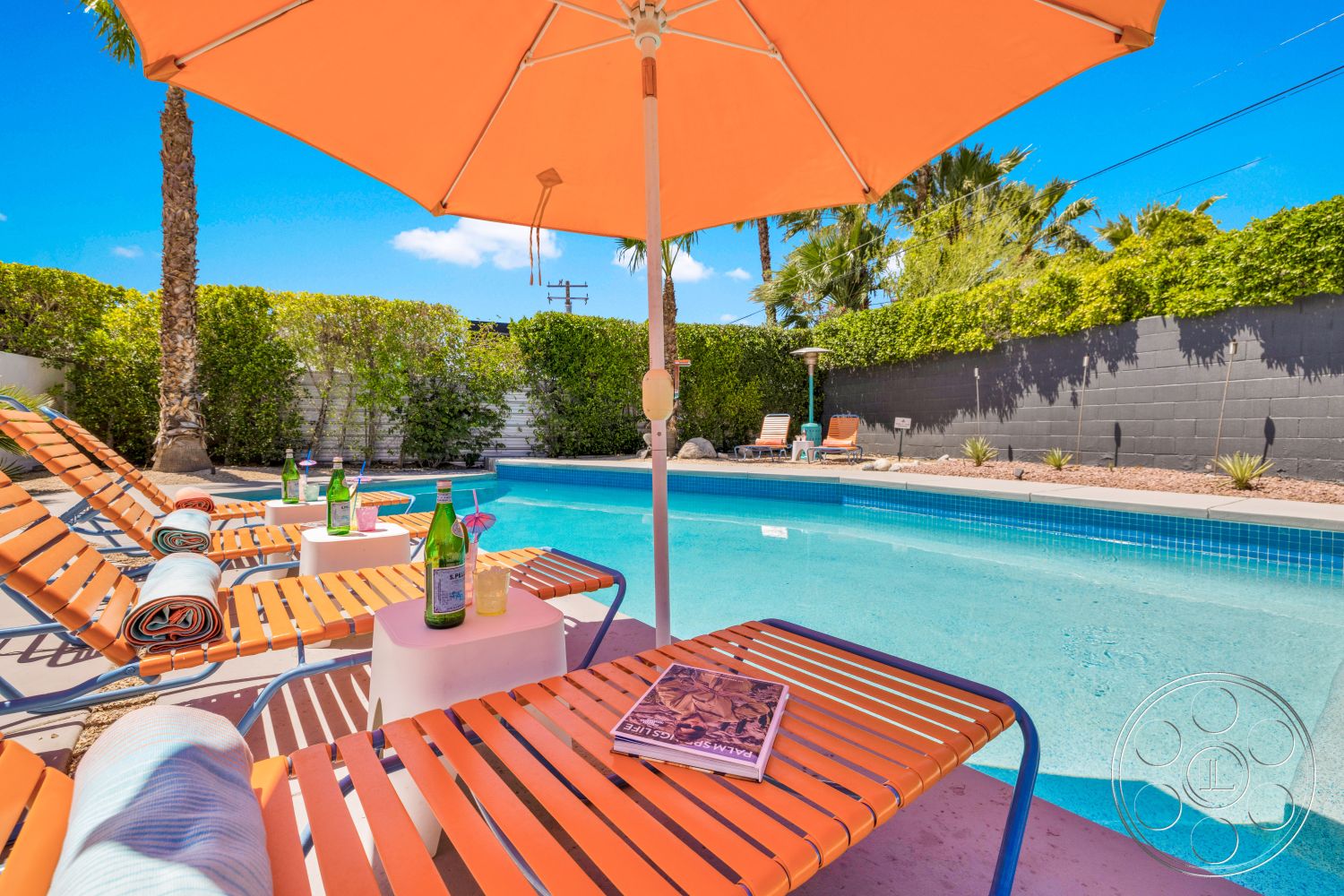 Palm Springs 76 - exterior, palm trees, gray concrete wall, clear blue sky, concrete pool deck, rectangular pool, sunny outdoor setting, orange patio umbrella, green shrub landscaping, tile pool border, orange metal lounge chairs, white outdoor side tables, beach towels, desert backyard