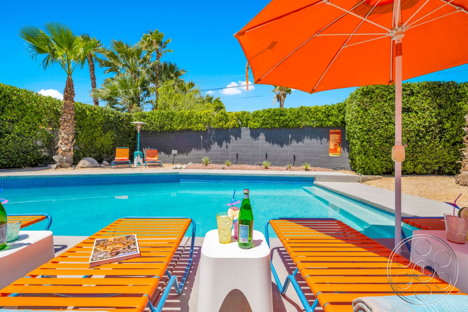 Palm Springs 76 - exterior, palm trees, blue sky, concrete pool deck, rectangular pool, sunny outdoor setting, outdoor umbrella, green hedge landscaping, desert backyard, blue tiled pool edge, orange beach lounge chairs, white outdoor tables