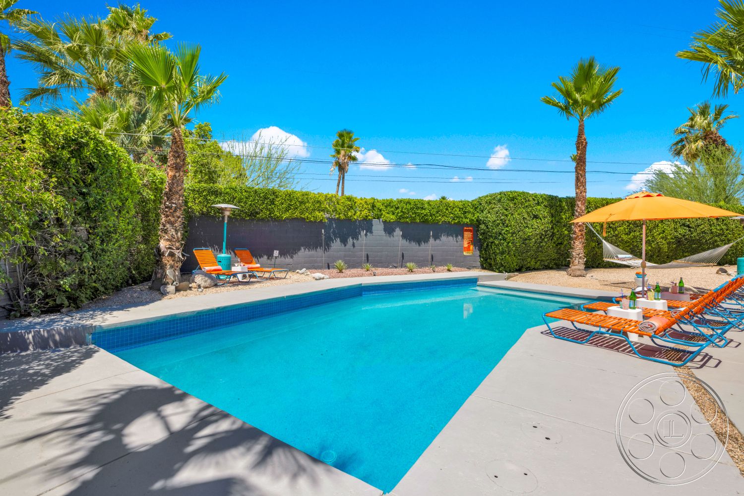 Palm Springs 76 - exterior, palm trees, rectangular pool, stone landscaping, hedge privacy wall, sunny outdoor setting, desert environment, concrete pool border, concrete pool patio, orange patio umbrella, blue mosaic tile pool, orange lounge chairs