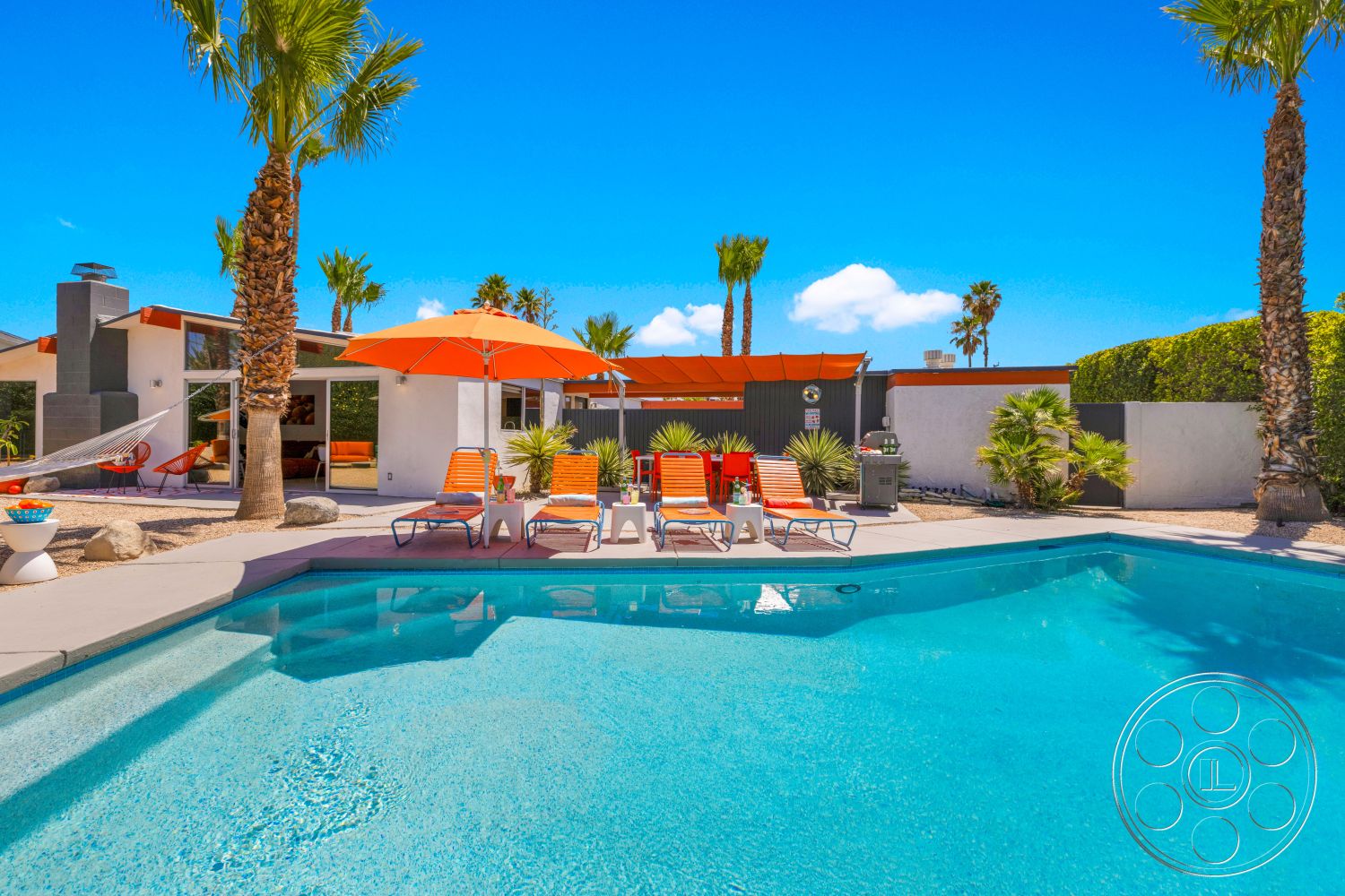 Palm Springs 76 - exterior, palm trees, glass sliding doors, outdoor dining area, outdoor lounge area, concrete pool deck, rectangular pool, minimalist landscaping, desert setting, stone landscaping, mid-century modern architecture, modern home design, sunny sky, orange patio umbrellas, hammock area, colorful pool chairs, grey stone wall
