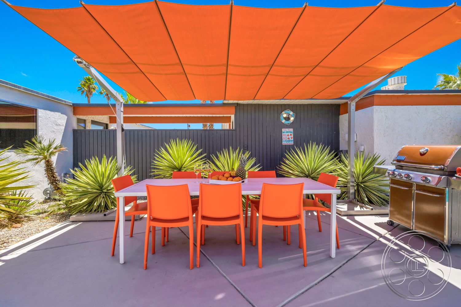 Palm Springs 76 - exterior, outdoor dining area, concrete patio flooring, blue sky backdrop, orange fabric shade canopy, modern white dining table, bright orange plastic dining chairs, built-in stainless steel grill, landscaped with desert plants, gray wooden fence
