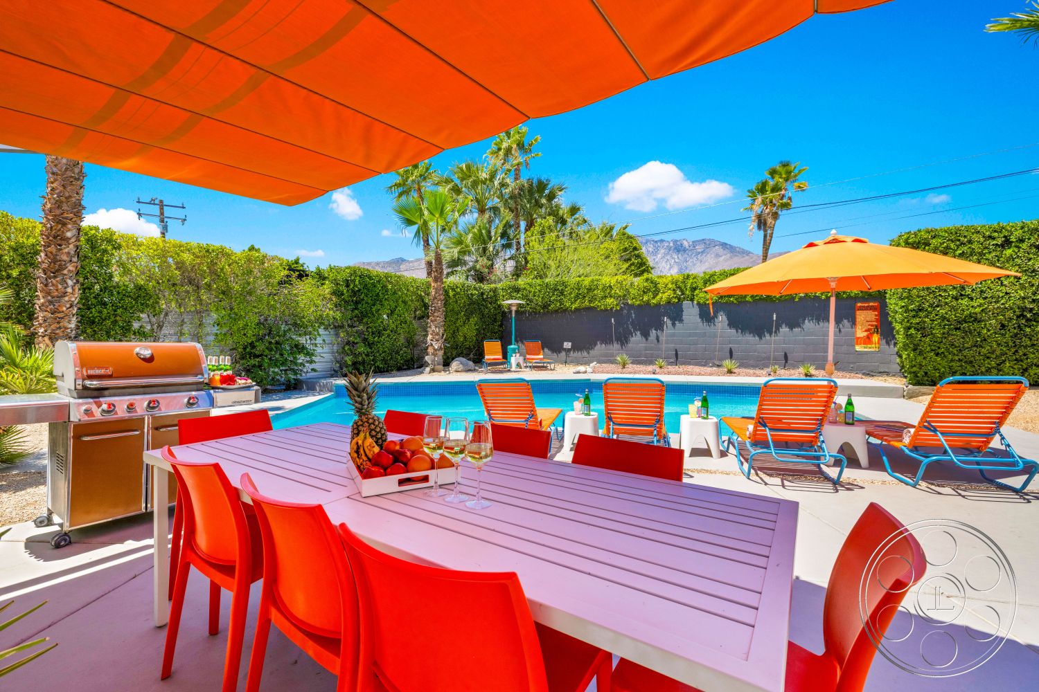 Palm Springs 76 - landscaped garden, palm trees, outdoor patio, rectangular swimming pool, mountain view, concrete pool deck, hedge privacy wall, white wooden dining table, orange patio umbrellas, steel outdoor grill, orange plastic dining chairs