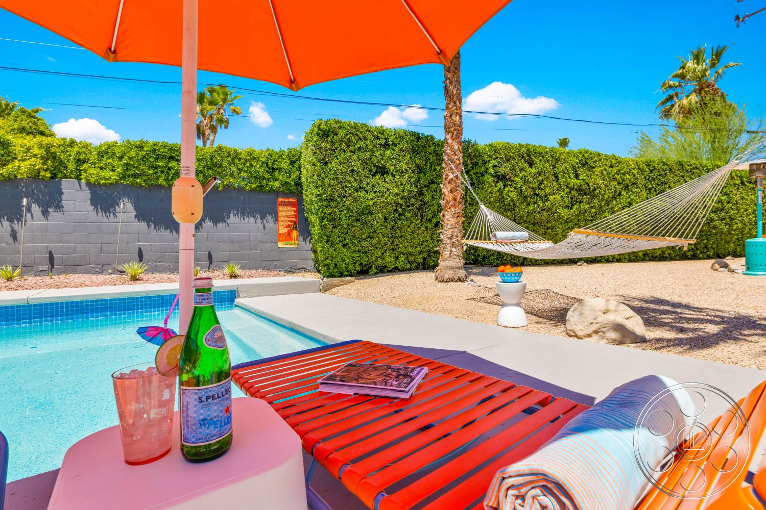Palm Springs 76 - outdoor space, rectangular swimming pool, casual seating area, blue sky, tropical landscaping, concrete pool deck, decorative stone, hammock, wooden lounge chair, sunny exterior, pedestal table, orange umbrella, fully landscaped backyard, green hedge wall